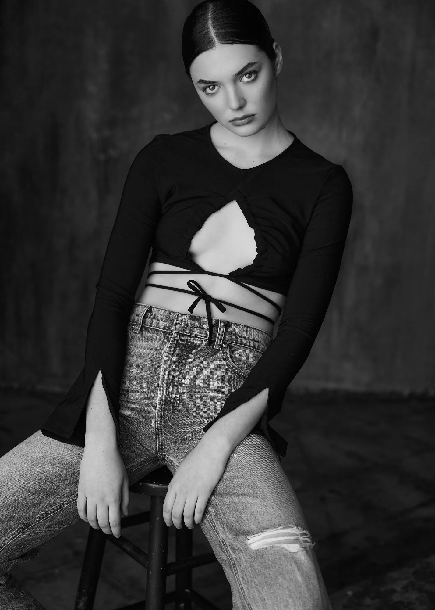 A black and white photo of a young woman with dark hair pulled back, wearing a black long-sleeve top with cutouts and high-waisted, ripped jeans, seated on a chair against a dark background.