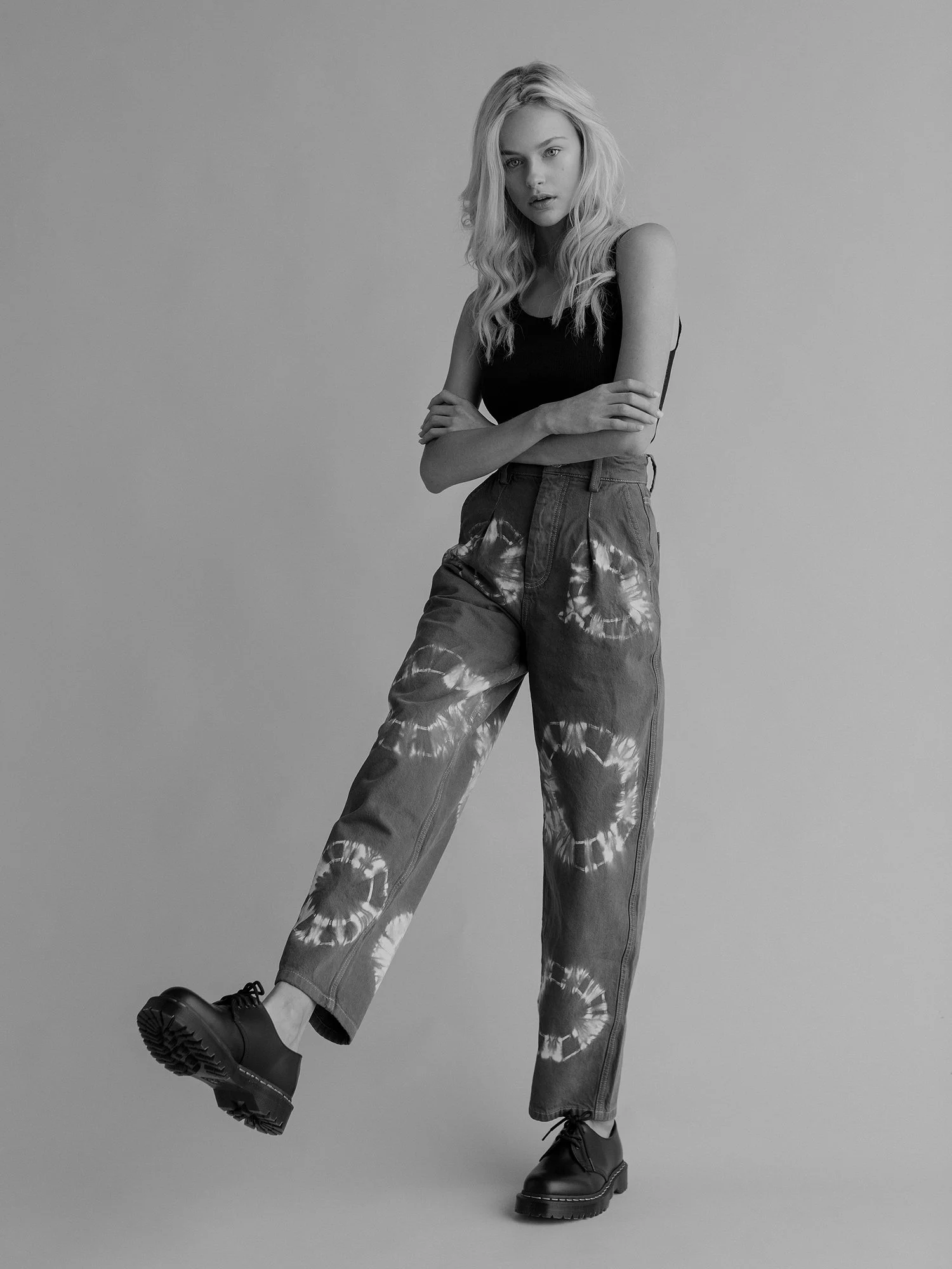 Black and white photo of a young woman with long wavy hair, wearing a sleeveless top, tie-dye pants, and chunky boots, standing against plain background, with one leg lifted and arms crossed.