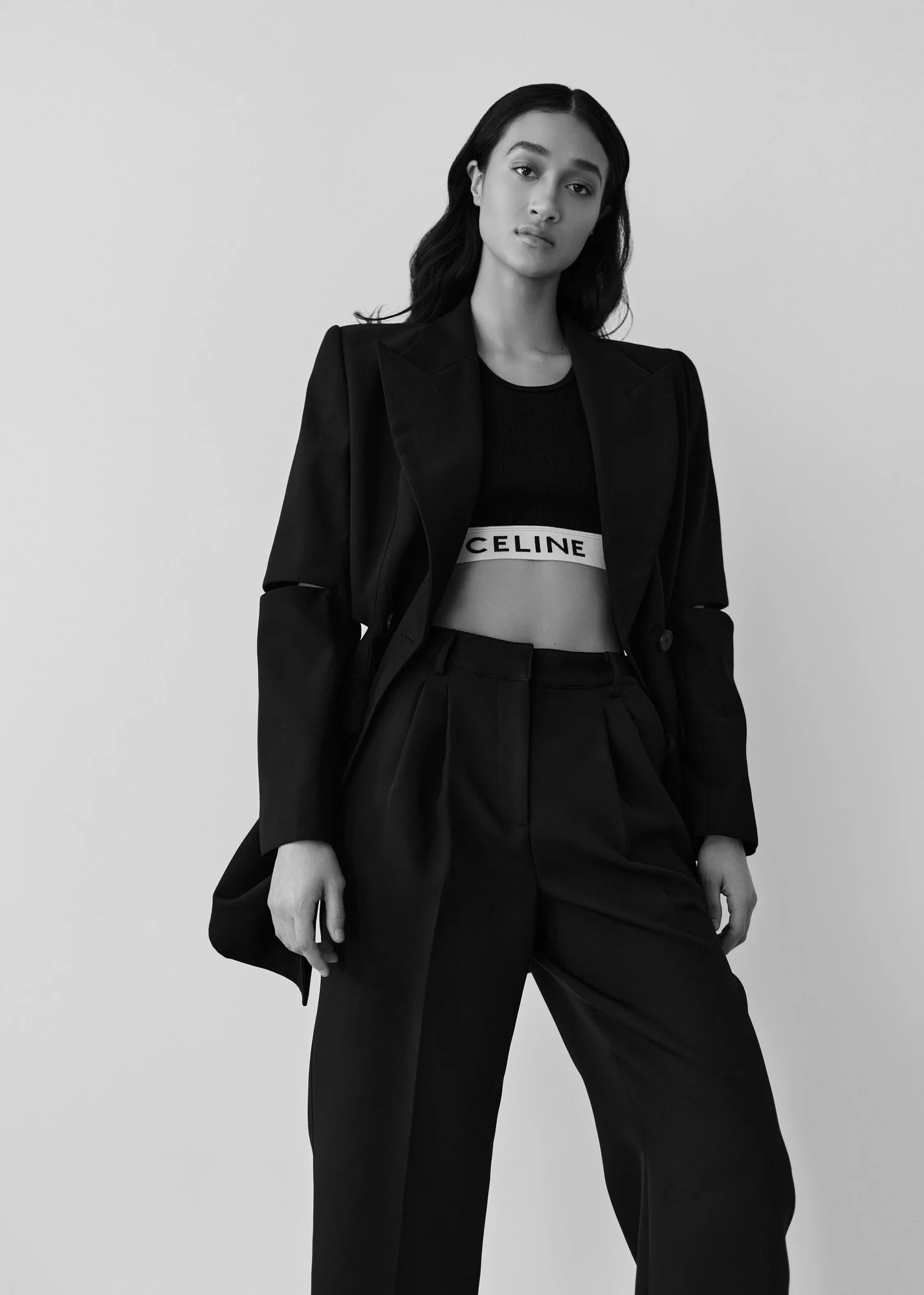 A woman wearing a black blazer and high-waisted black pants, with a black crop top displaying the brand name 'CELINE,' standing against a plain light background.