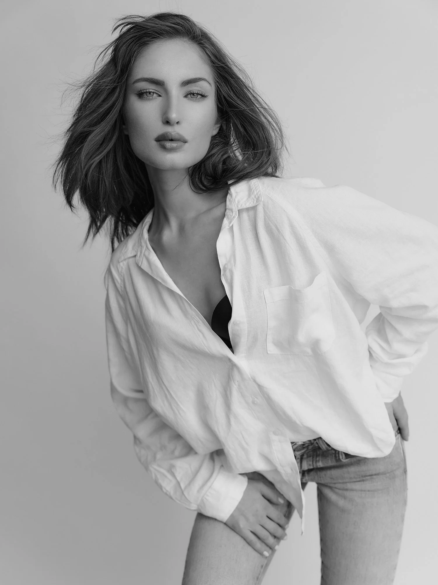 Black and white photograph of a woman with shoulder-length hair, wearing a loose white shirt and jeans, posing with her hand on her thigh against a plain light background.