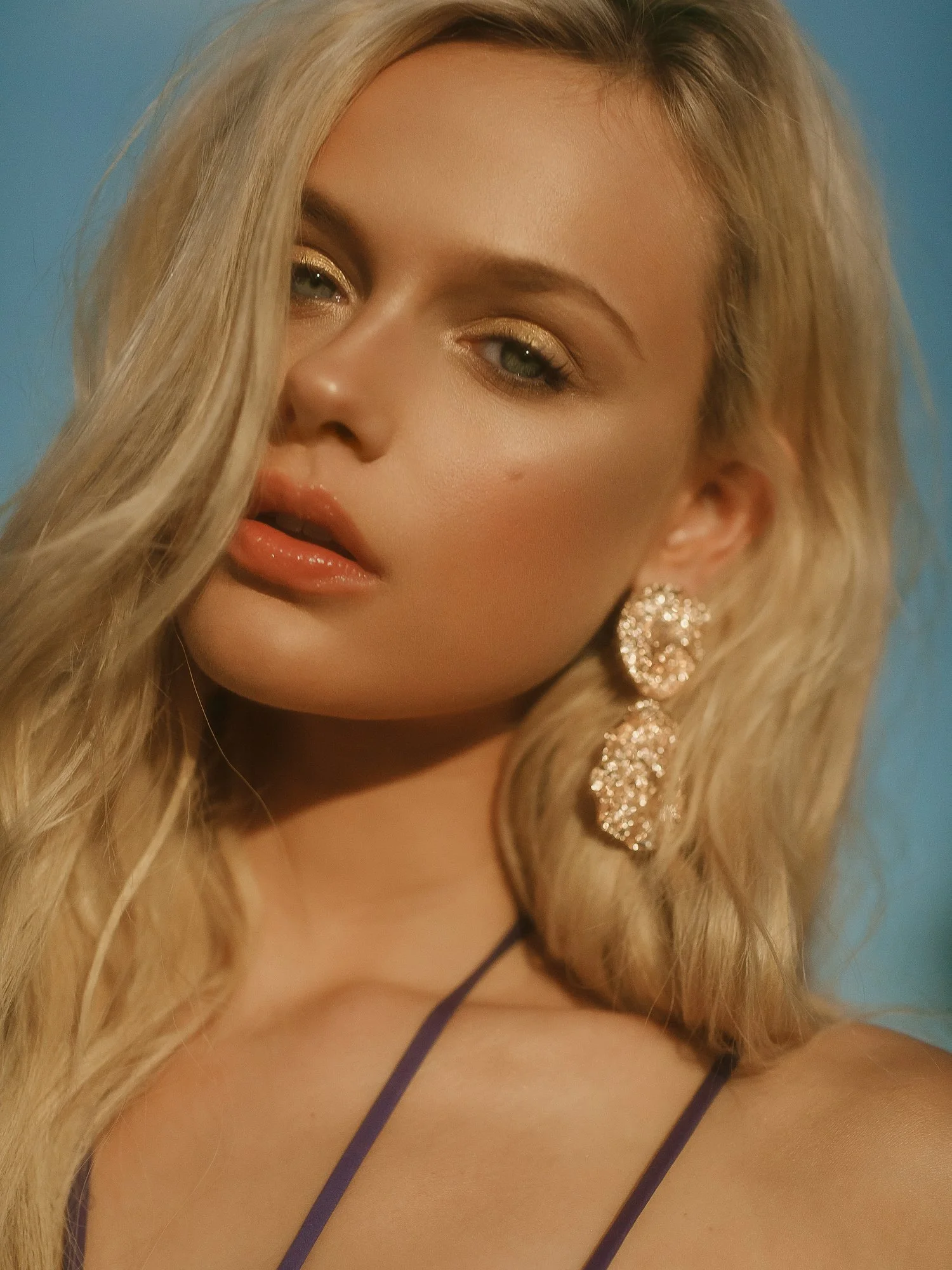 Close-up of a woman's face with blonde hair, gold eyeshadow, and large sparkling earrings, against a blue background.