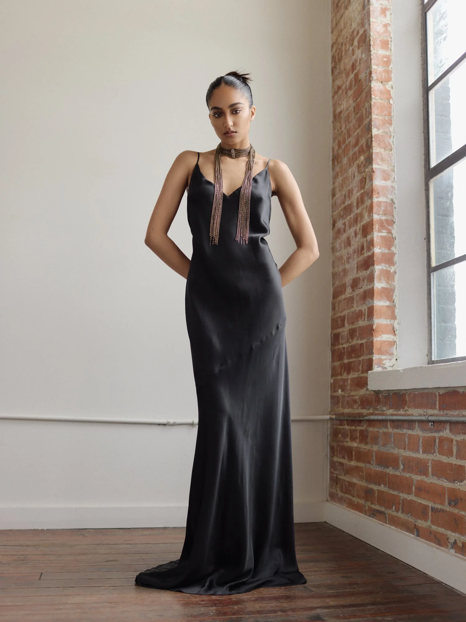 A woman wearing a black satin evening gown with spaghetti straps and a dramatic necklace, standing in a room with a white wall, a brick wall, and a large window with black frames.