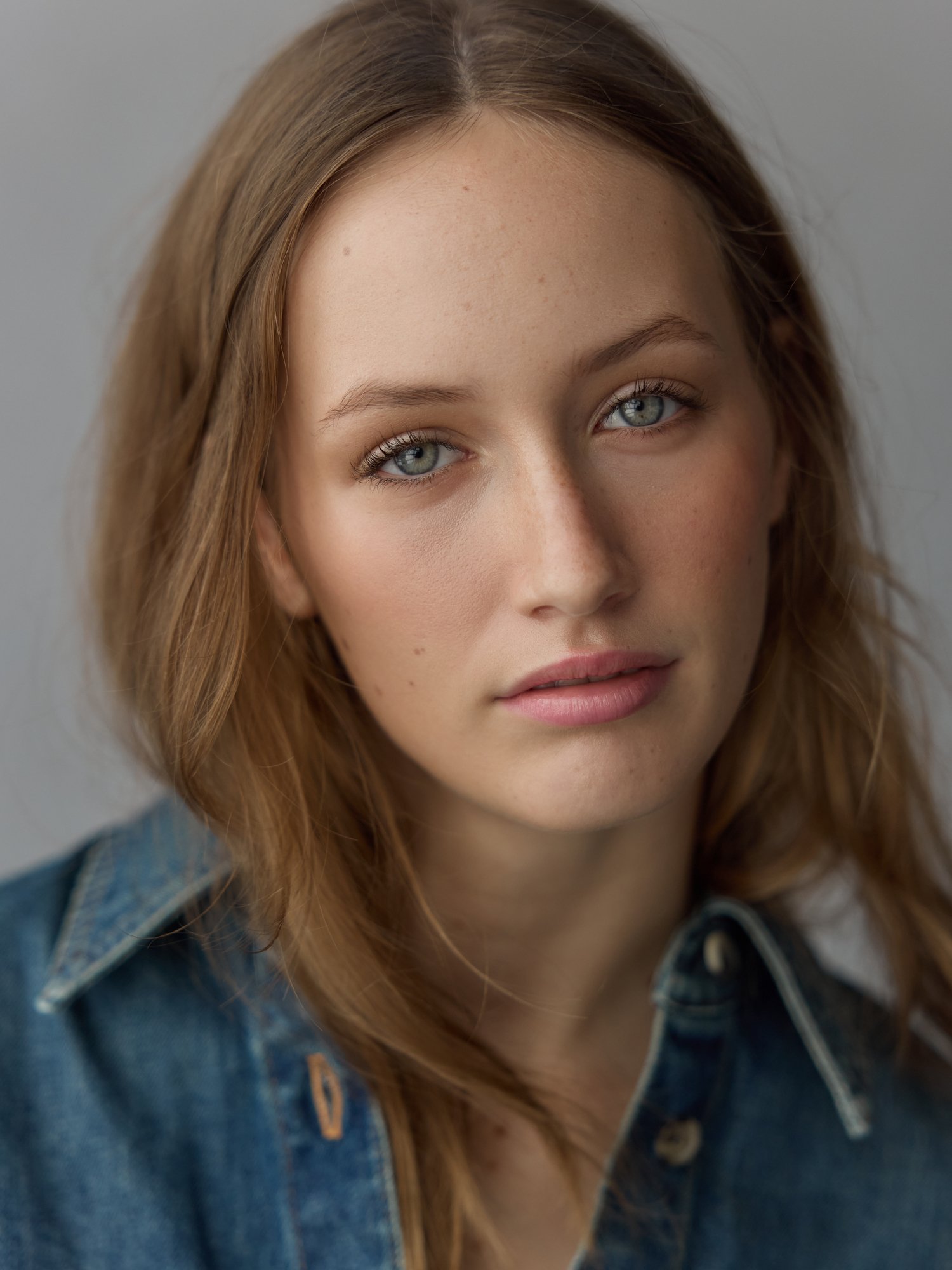 Close-up of a woman with long, light brown hair, blue eyes, and natural makeup wearing a denim jacket with a black shirt underneath.