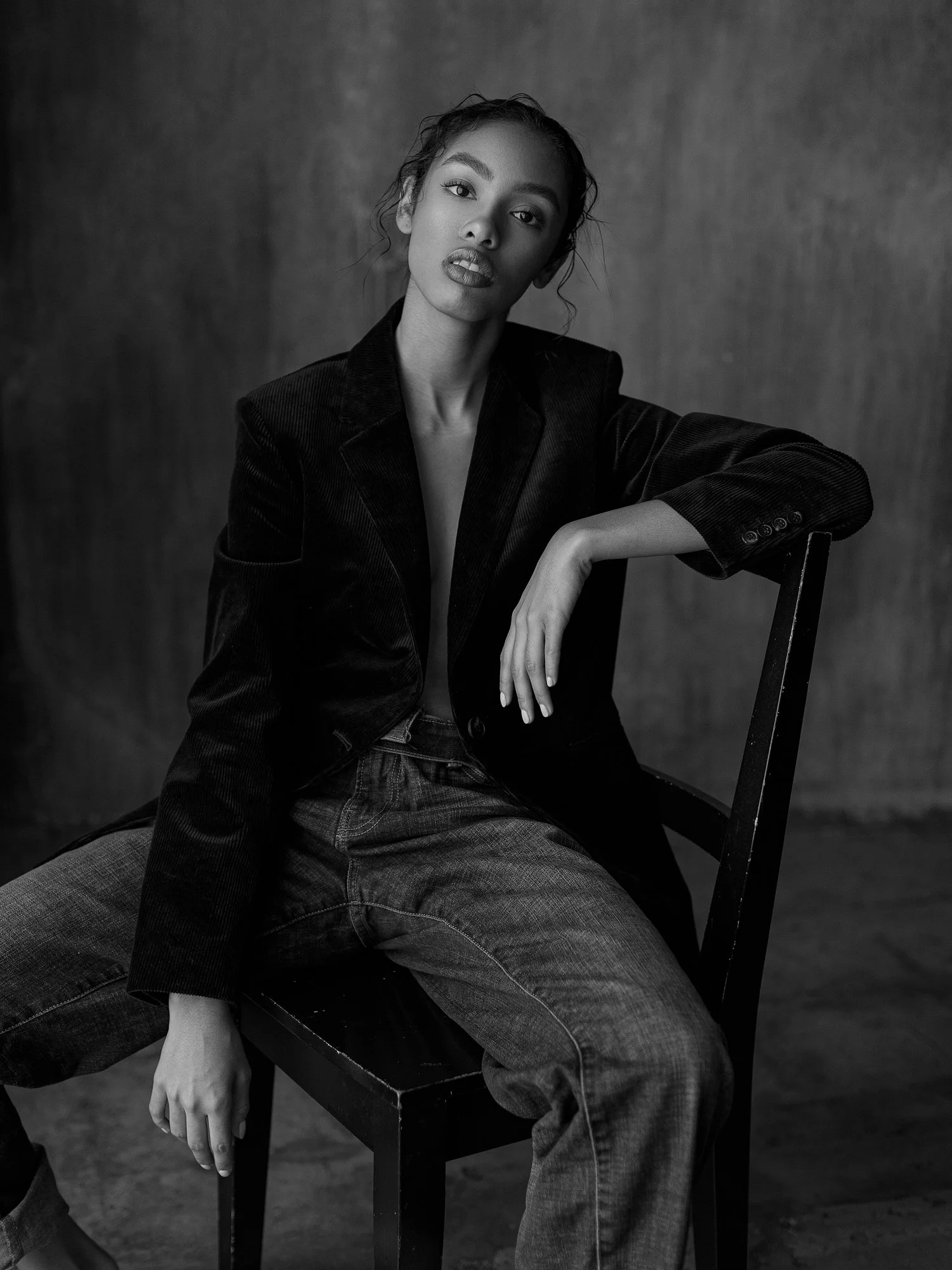 Black and white portrait of a woman sitting on a chair, wearing a dark blazer and jeans, with her arm resting on the back of the chair, gazing at the camera with a serious expression, against a dark plain background.