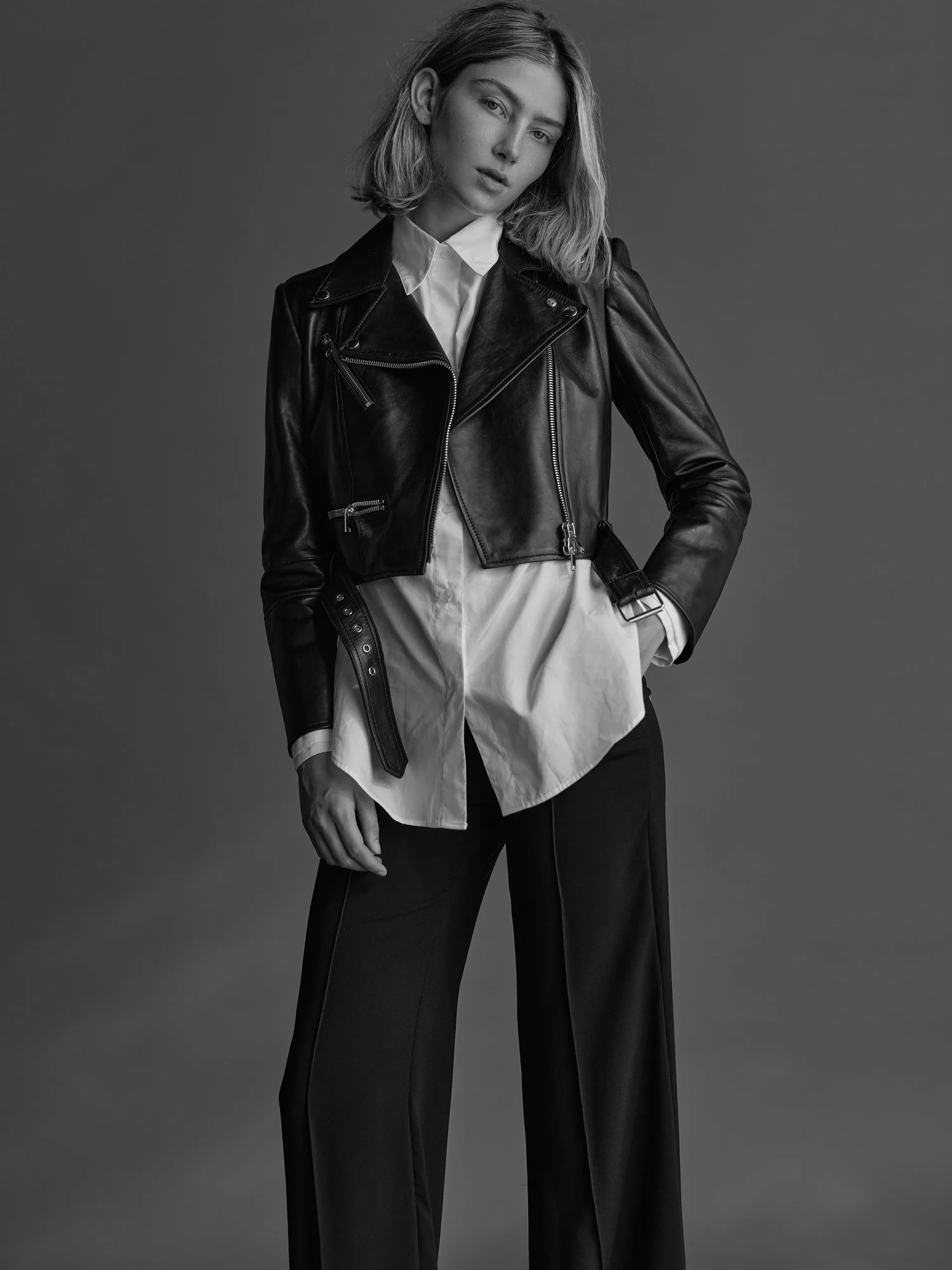 A young woman in a black leather jacket, white shirt, and black pants standing against a plain background.