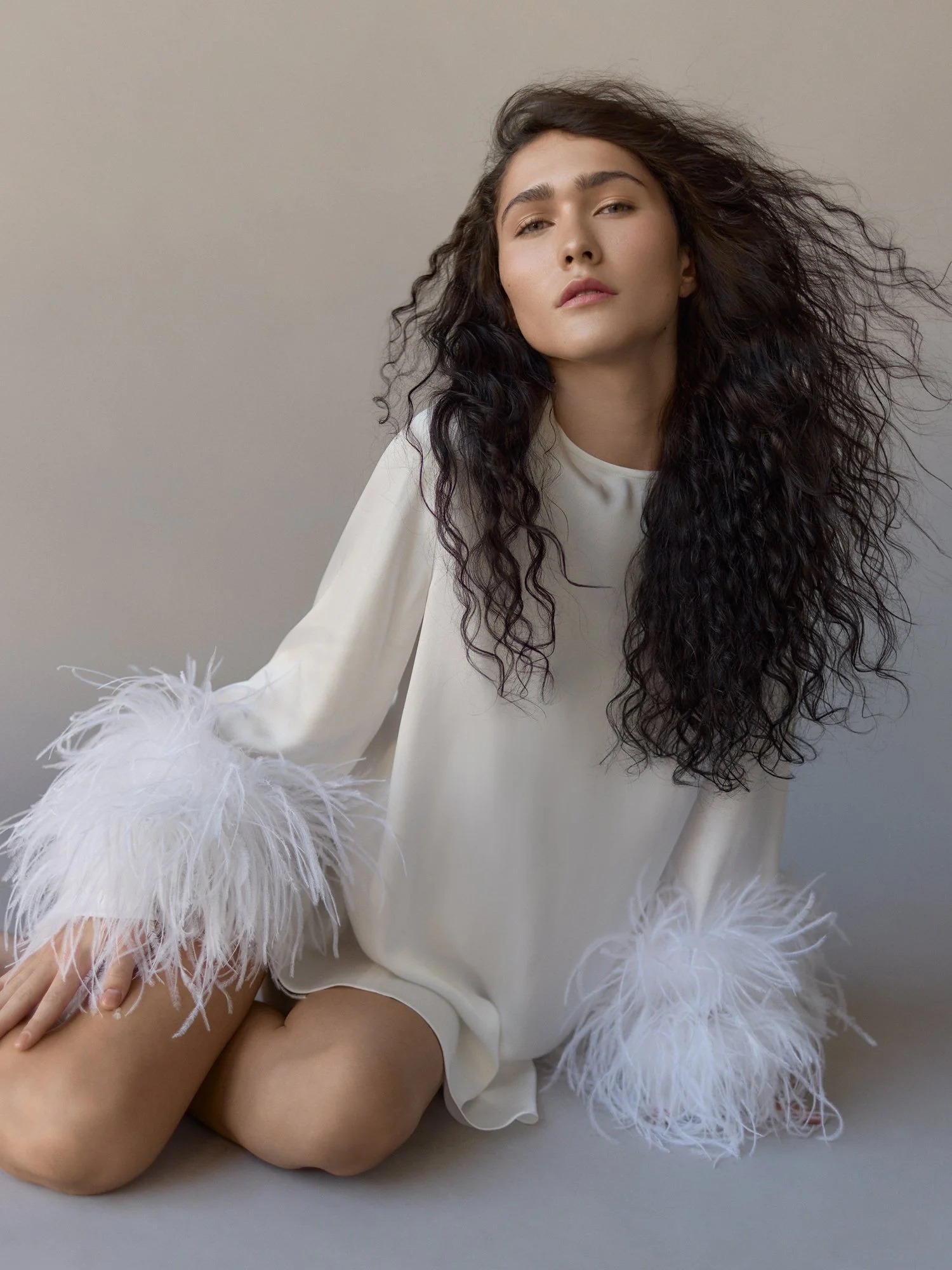 Woman in white satin robe with feather cuffs posing against a plain backdrop.