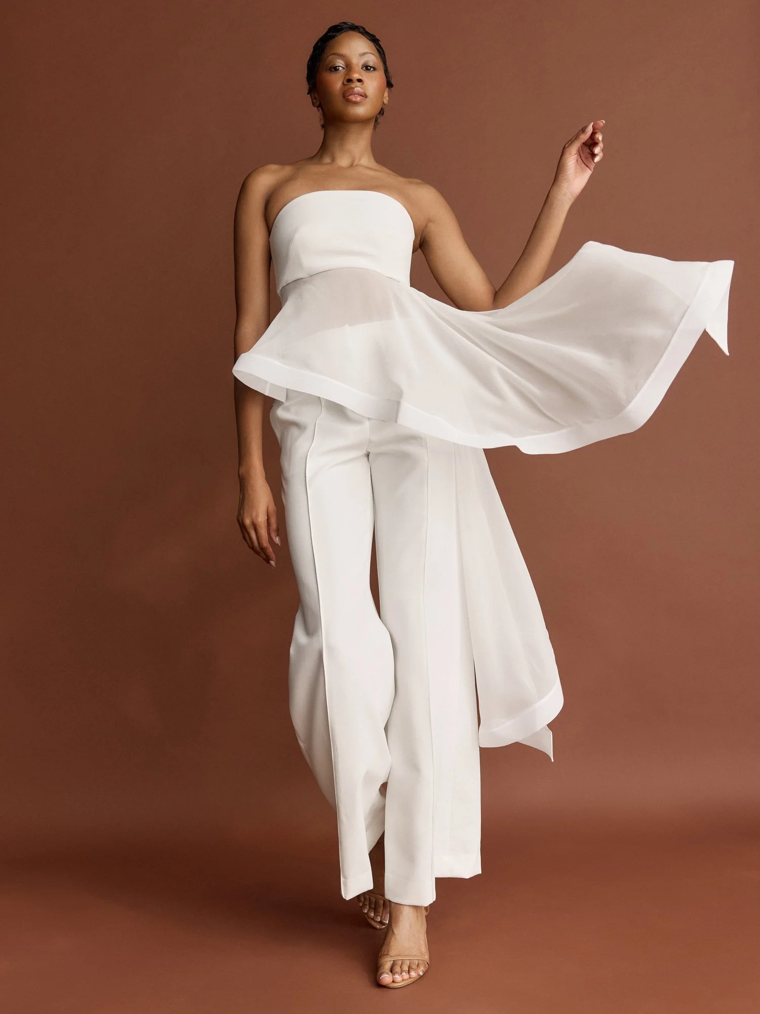 A woman with short hair wearing a strapless white top and wide-leg white pants, standing against a brown background, holding her left arm up with her hand slightly curled.