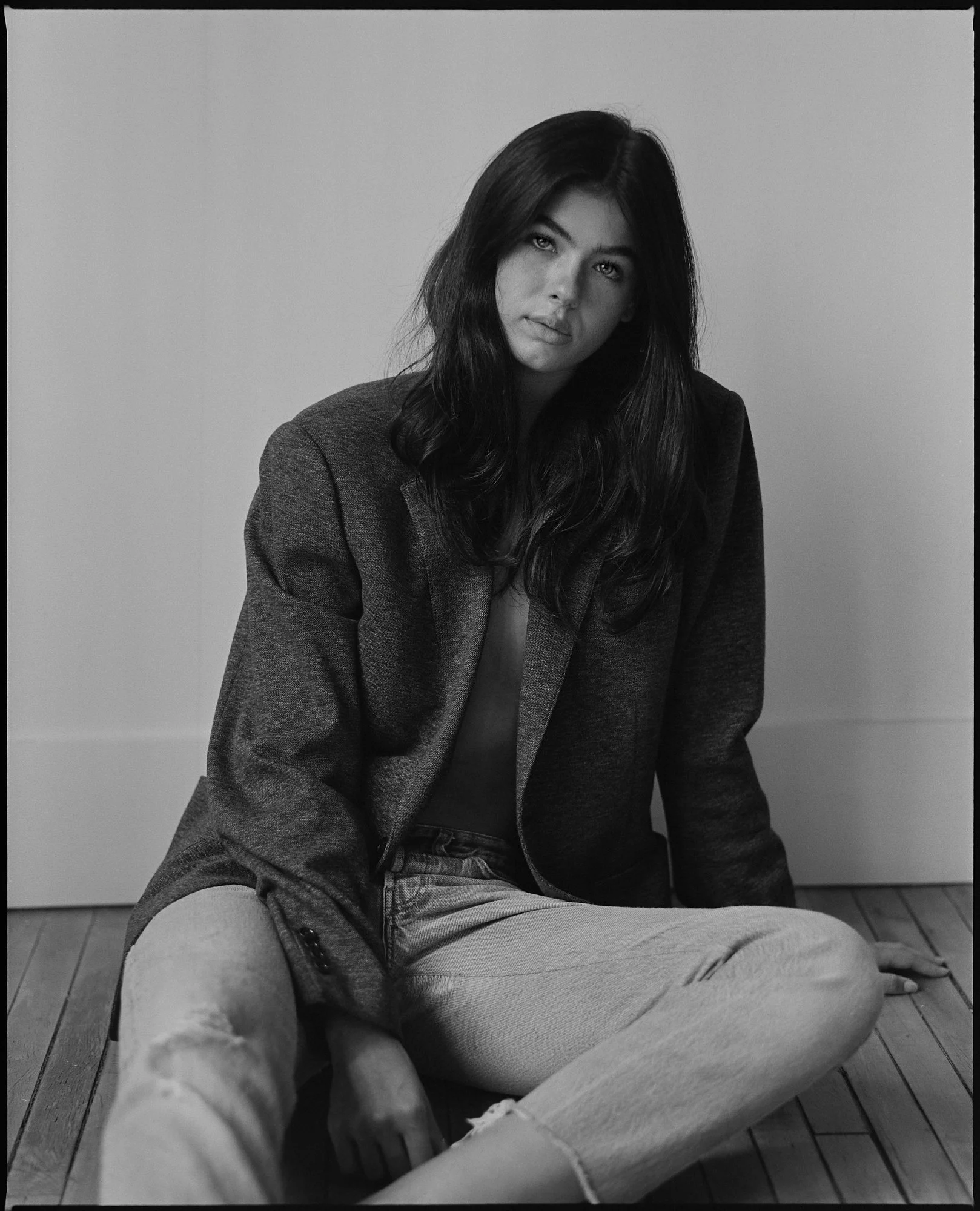Black and white photograph of a woman with long dark hair, dressed in a blazer and jeans, sitting on a wooden floor against a plain wall, looking at the camera with a neutral expression.