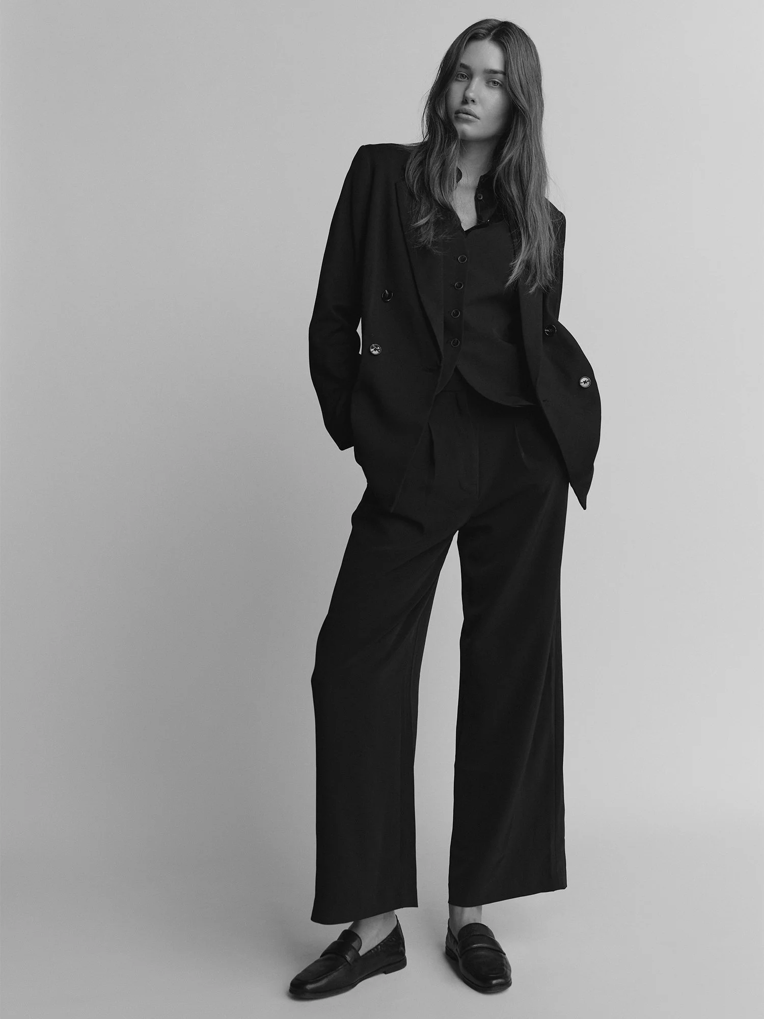 Black and white photo of a woman standing with her hands in her pockets, wearing a dark blazer, matching pants, and loafers, against a plain background.