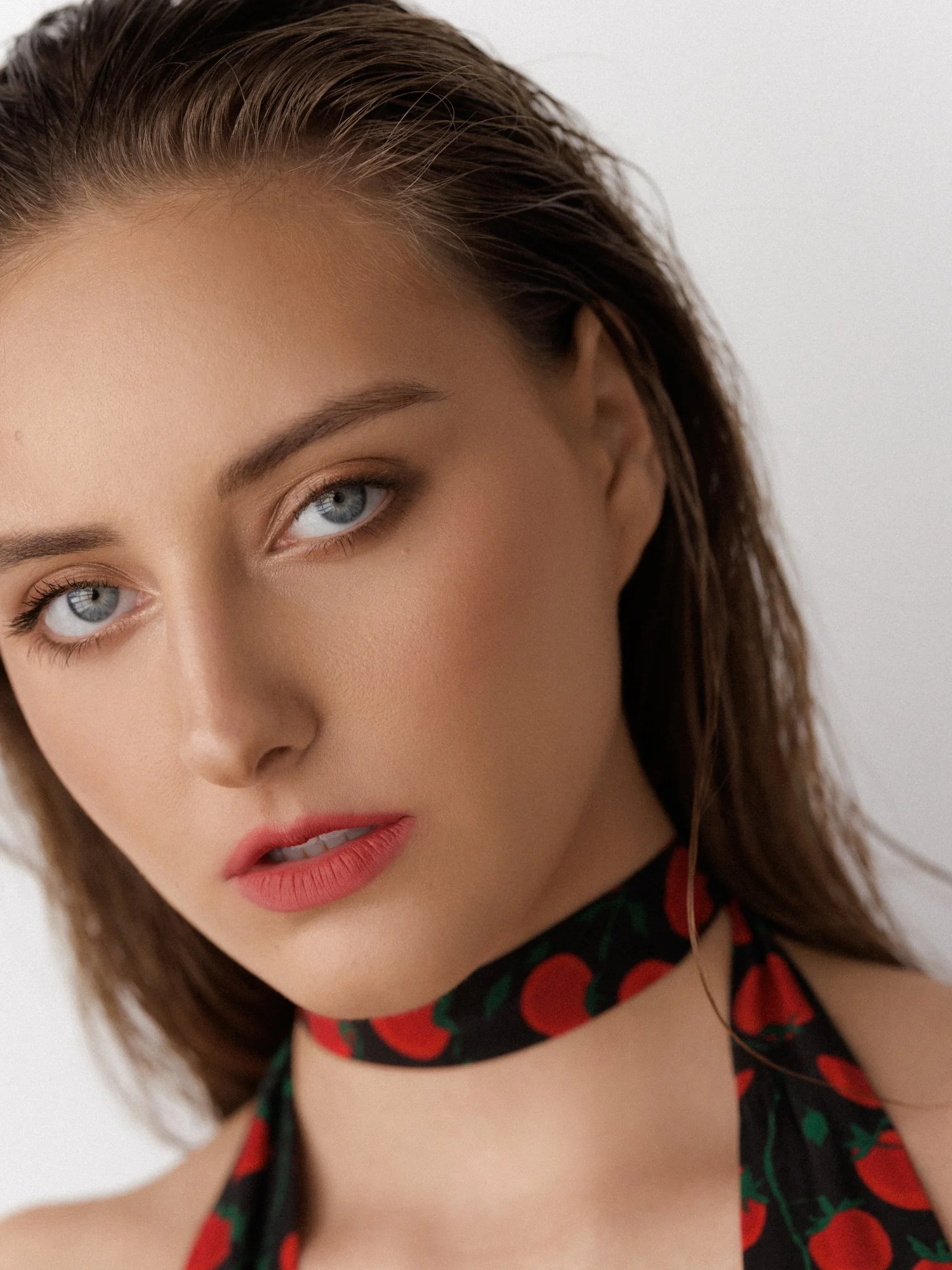 Close-up of a woman with blue eyes and brown hair, wearing red lipstick and a black dress with red cherries and green leaves, against a white background.