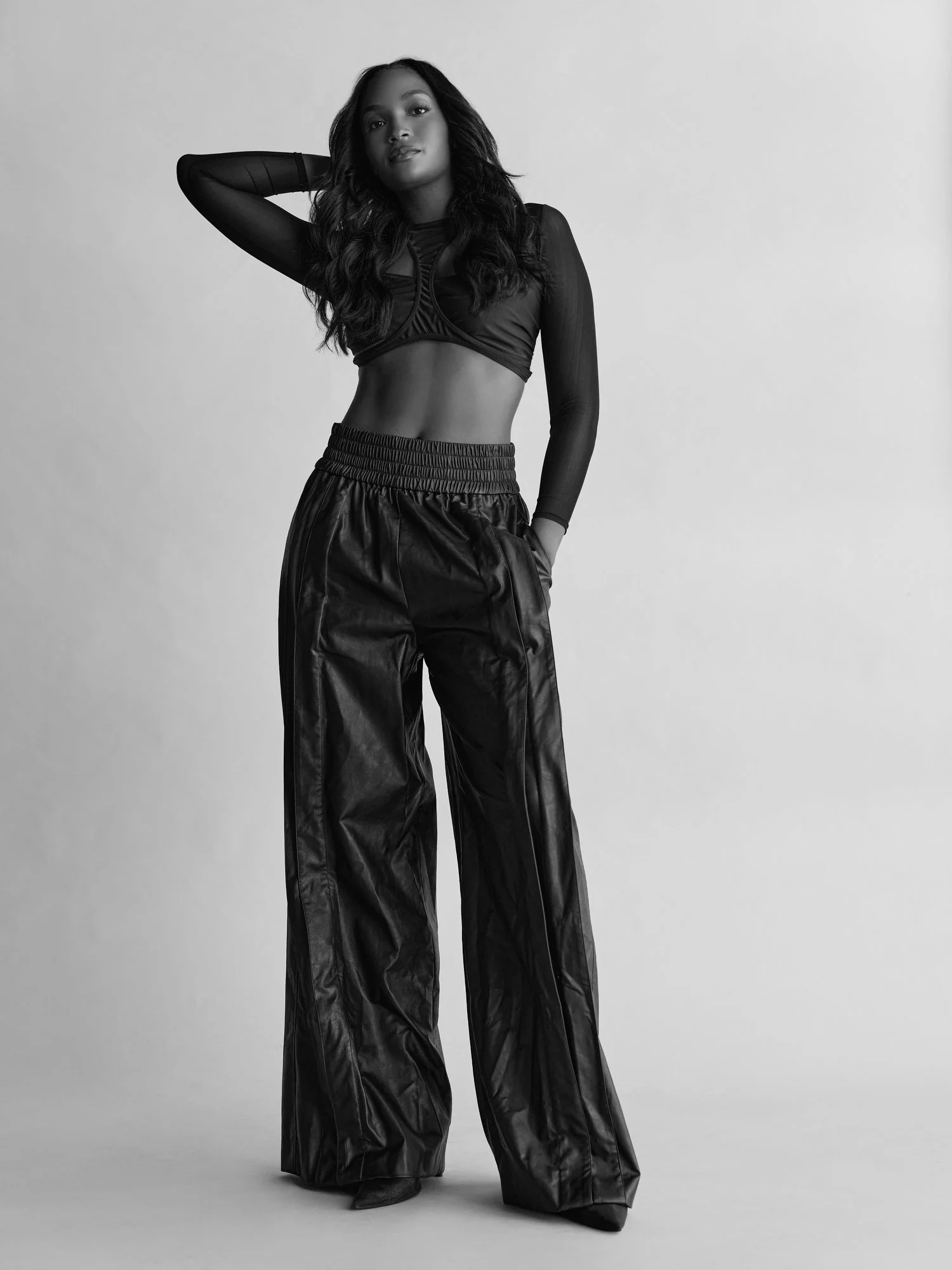A woman with long dark hair posing with one arm behind her head, wearing a black sheer long sleeve top and shiny black wide-leg pants, standing against a plain light gray background.