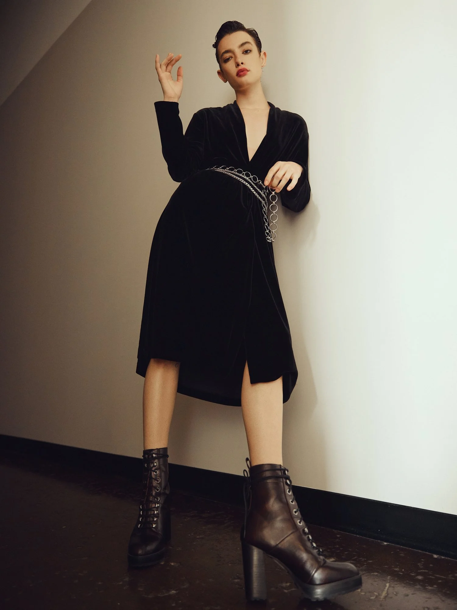 Young woman in black Velvet dress and brown lace-up platform boots standing against a plain wall, with one hand raised and the other on her hip.
