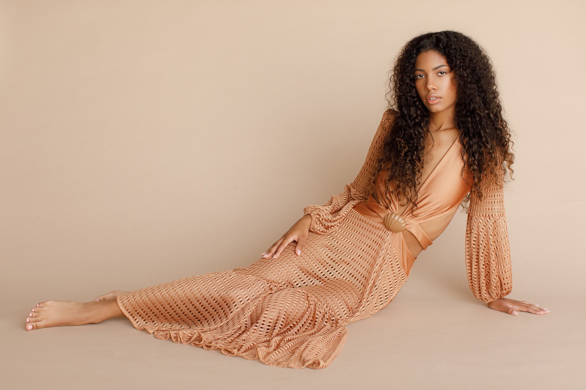 A woman with long curly hair, wearing a peach-colored crochet dress, sitting on the floor against a beige background.