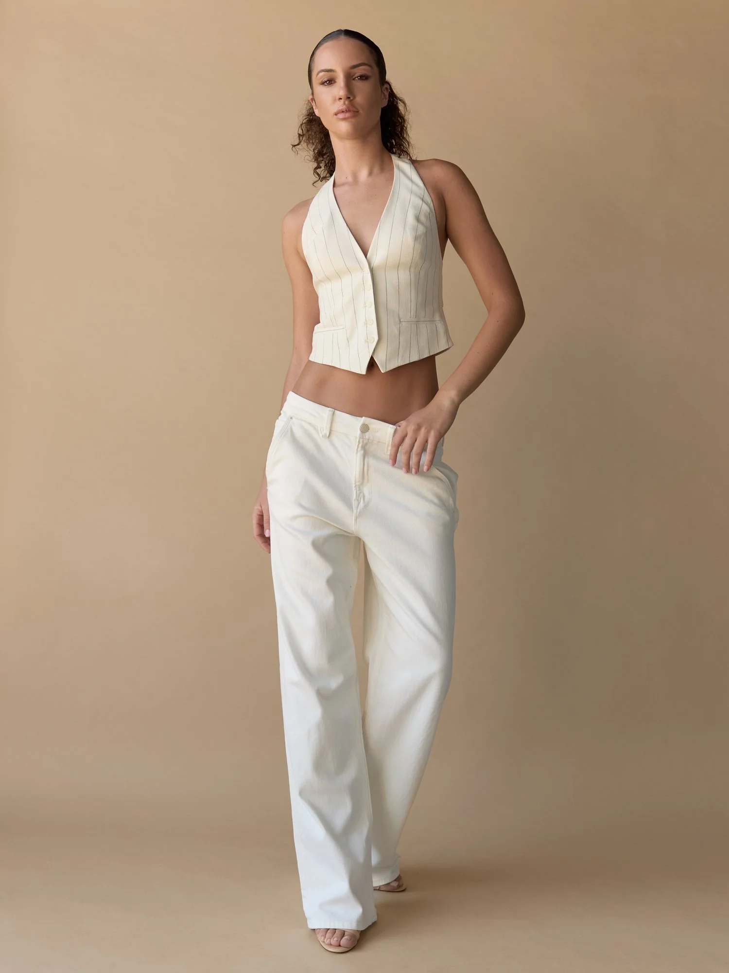 A woman wearing a sleeveless, cream-colored striped vest and loose white pants stands against a plain beige background.
