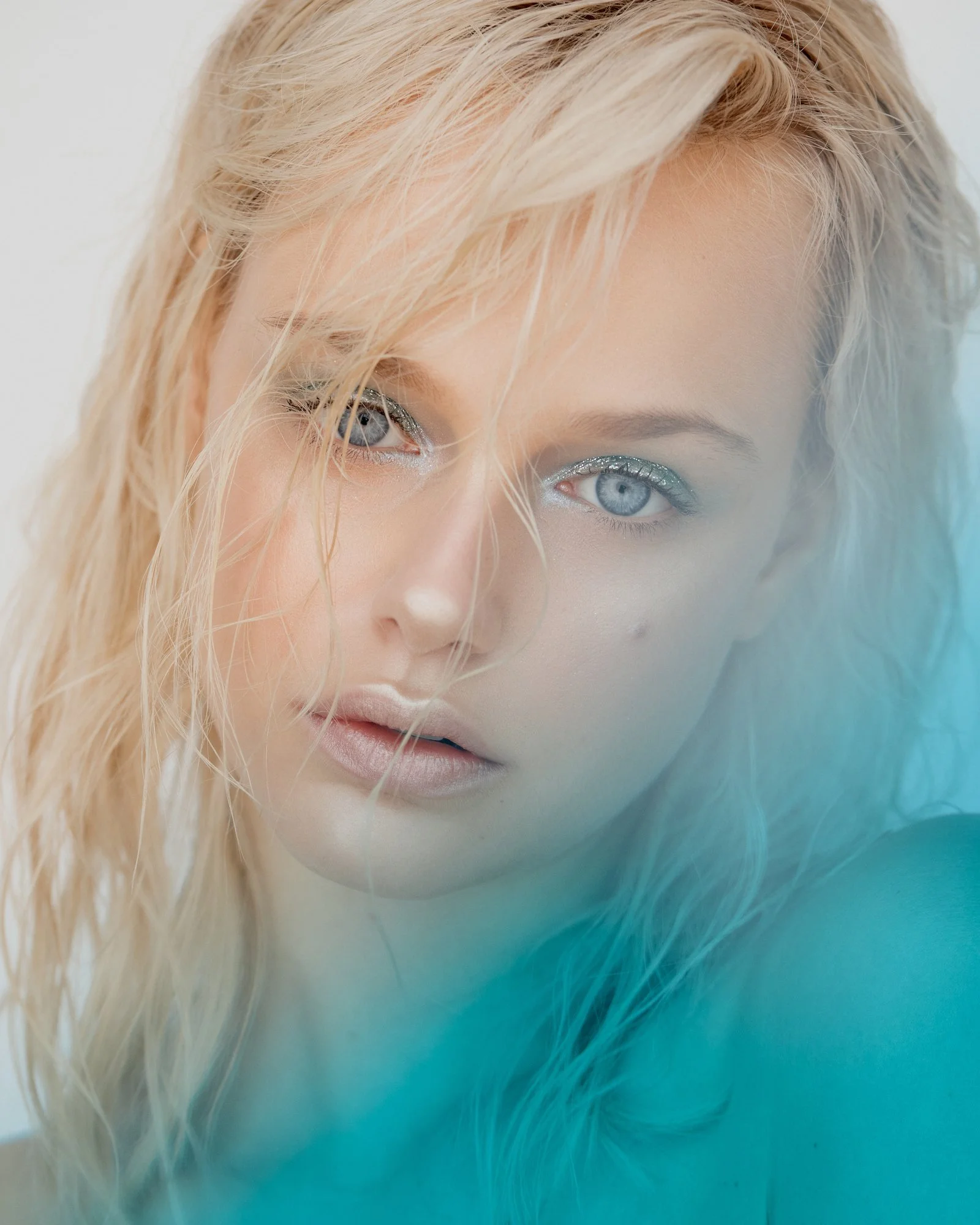 Close-up of a woman's face with blonde hair and blue eyes, wearing silver glitter eye makeup, against a blurred background with a gradient of light blue and teal colors.