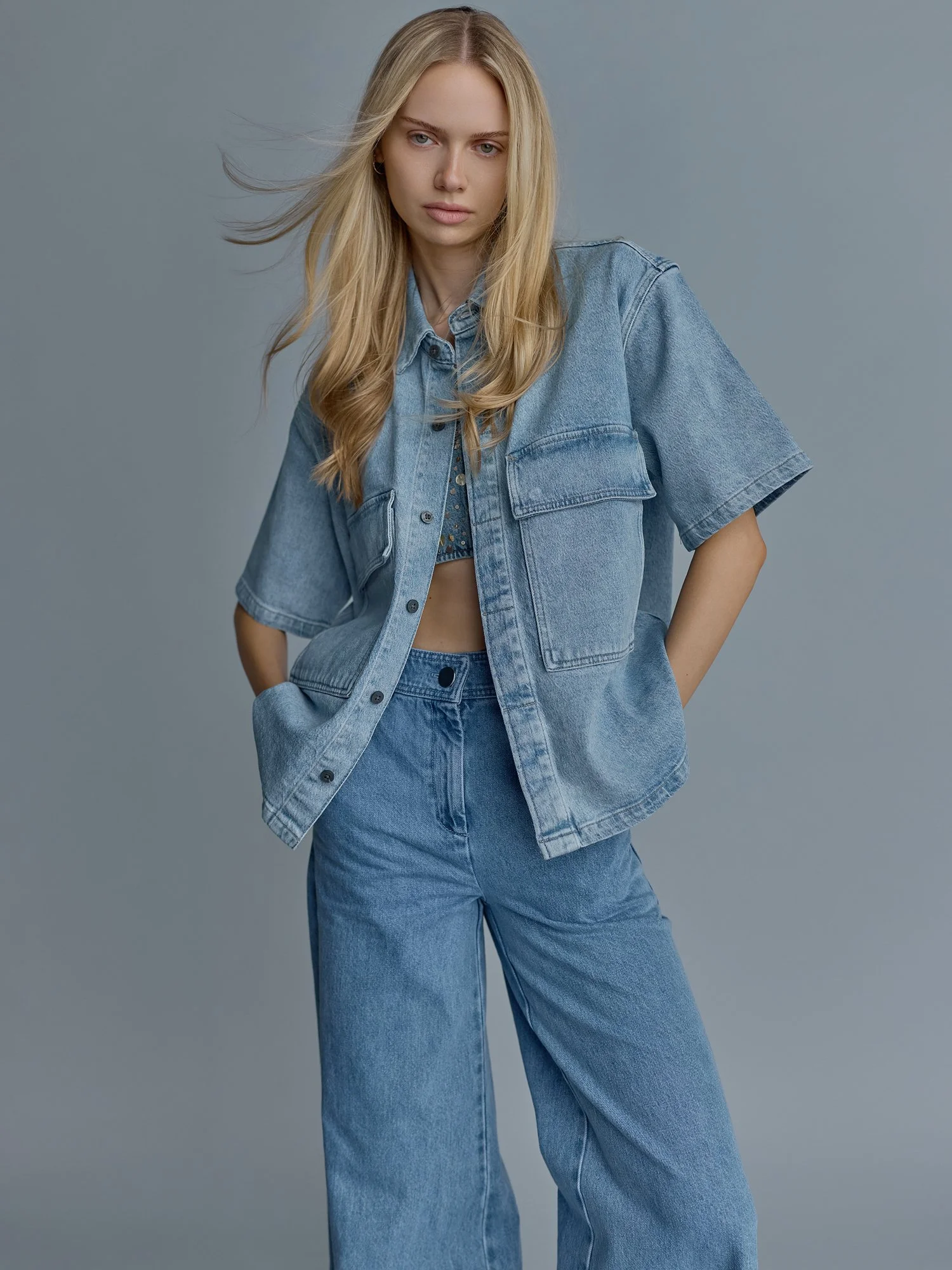 A young woman with long blonde hair wearing a denim jacket and matching high-waisted jeans against a plain gray background.