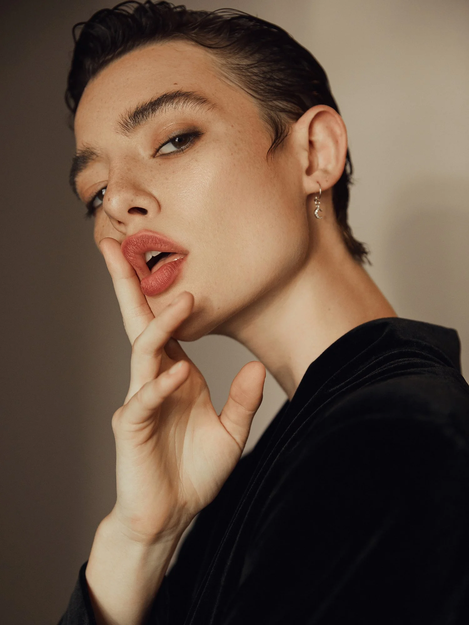 Close-up of a woman with dark, short hair and freckles, wearing a black top and earrings, touching her lips with her fingers.