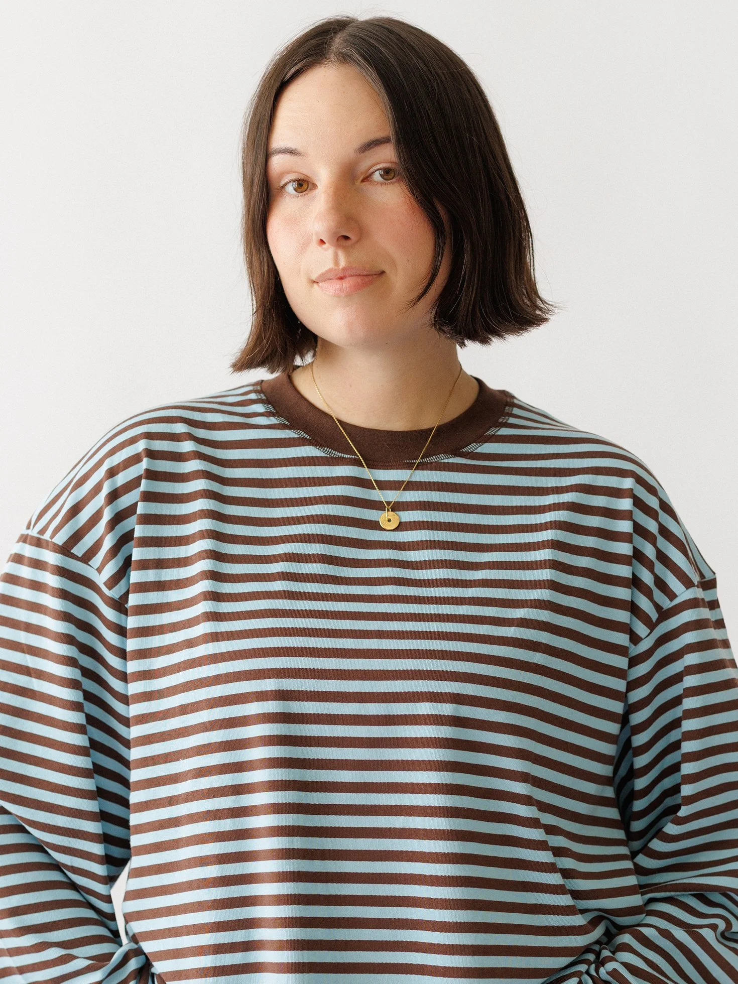 A woman with shoulder-length dark hair in a brown and light blue striped long-sleeve shirt, wearing a gold necklace, standing against a plain white background.