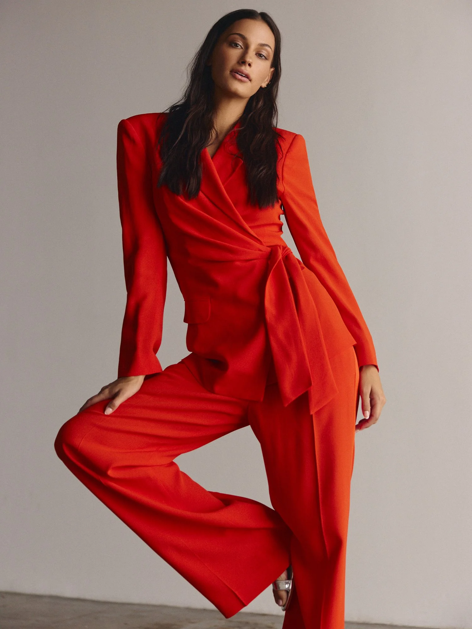 A woman wearing a red tailored blazer and red pants poses against a plain background. She has long dark hair and is wearing minimal makeup, with her hand resting on her bent knee.