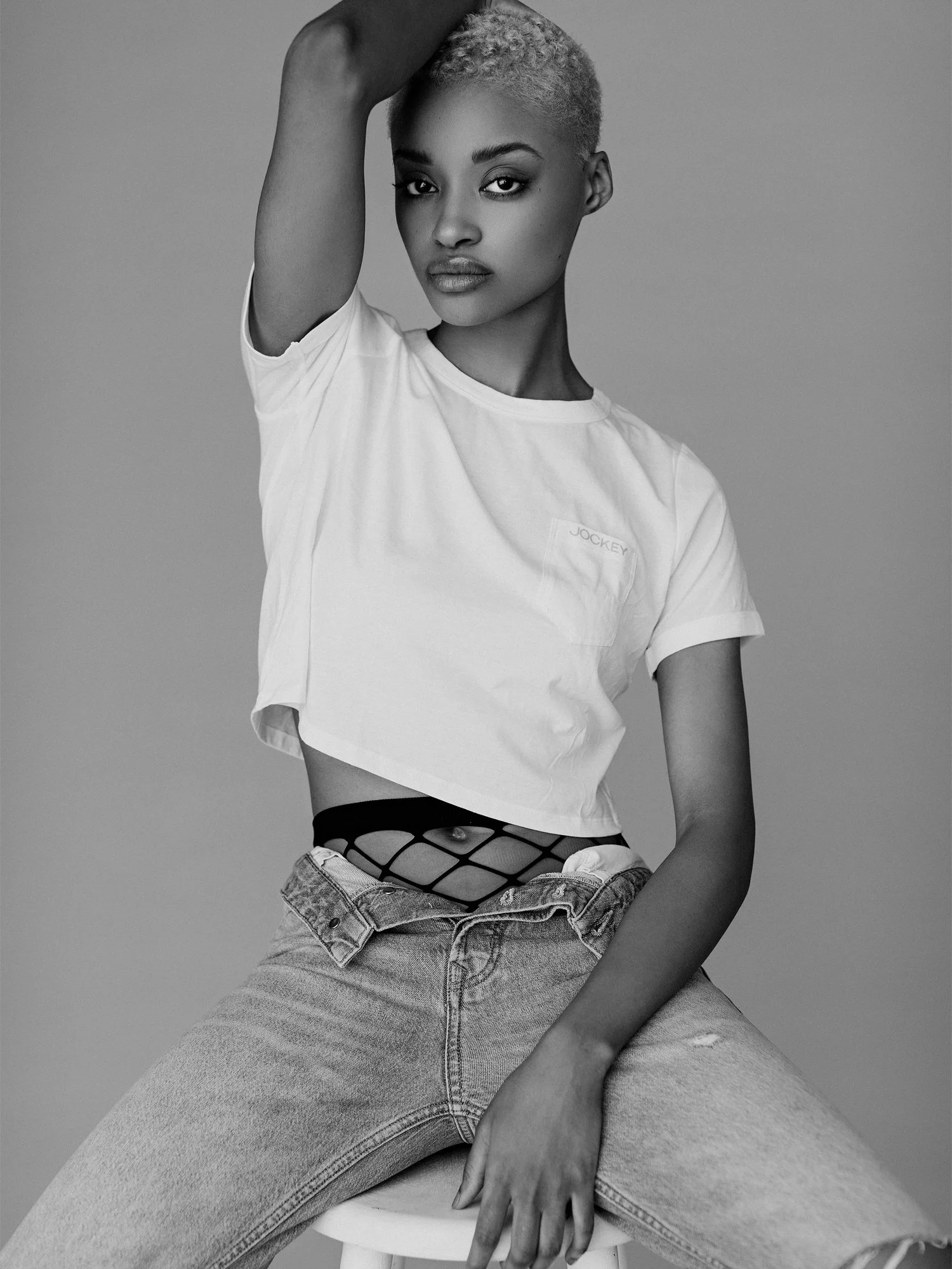 Black and white photograph of a young woman with short, curly blonde hair, wearing a loose white t-shirt with the brand name 'Jockey' on the chest, fishnet stockings under ripped jeans, and sitting on a white stool with her legs apart. She has an int