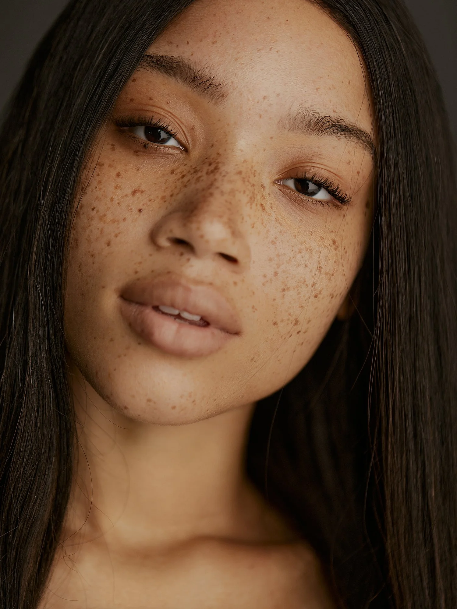 Close-up of a woman with long dark hair, brown eyes, and visible freckles on her face.