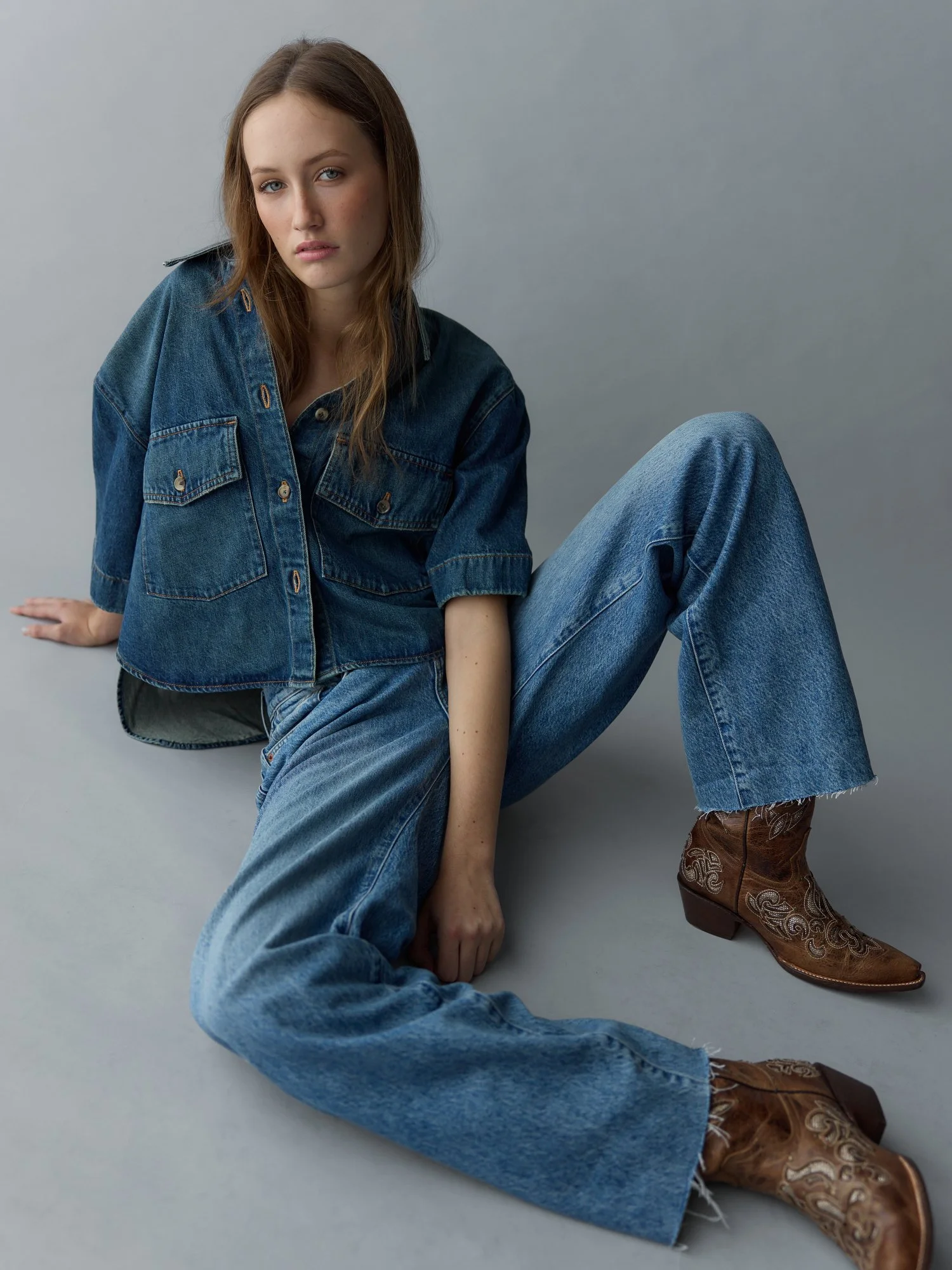 A young woman with light skin and long brown hair, wearing a denim jacket and jeans, sitting on the floor with one leg bent and the other stretched out, wearing cowboy boots, against a plain gray background.