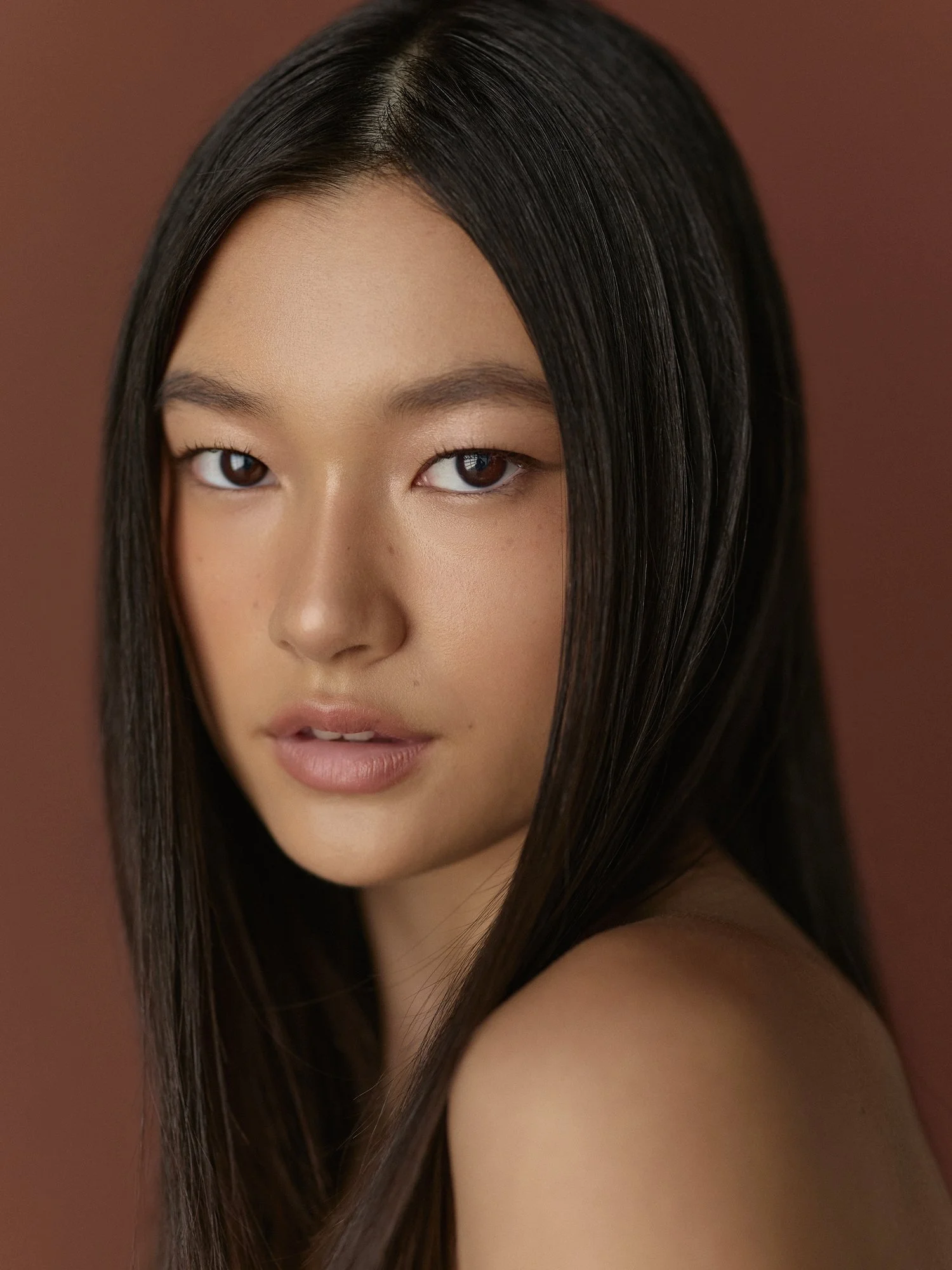 Close-up of a woman's face with long, straight black hair, neutral makeup, and a warm brown background.