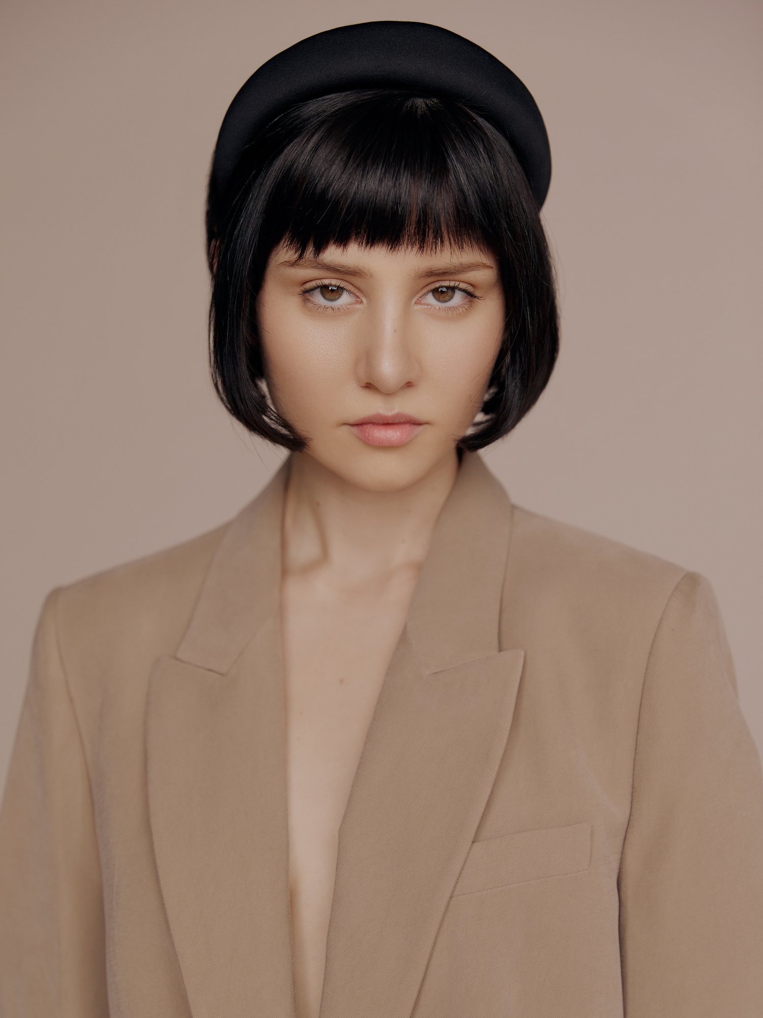 A woman with short black hair, wearing a beige blazer and a black beret, looking directly at the camera against a neutral background.