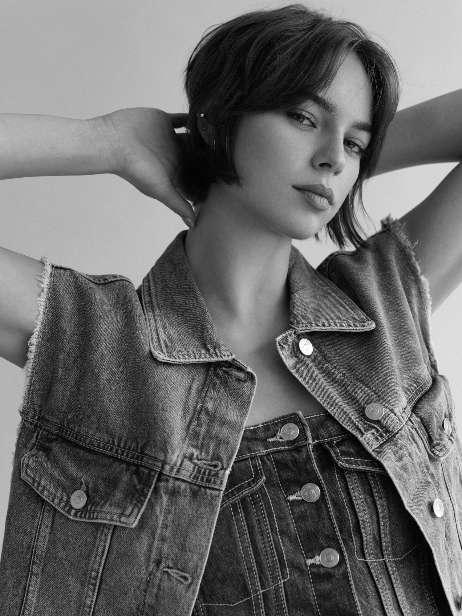 Black and white portrait of a woman with short hair, wearing a denim vest and a striped shirt, standing against a plain background.