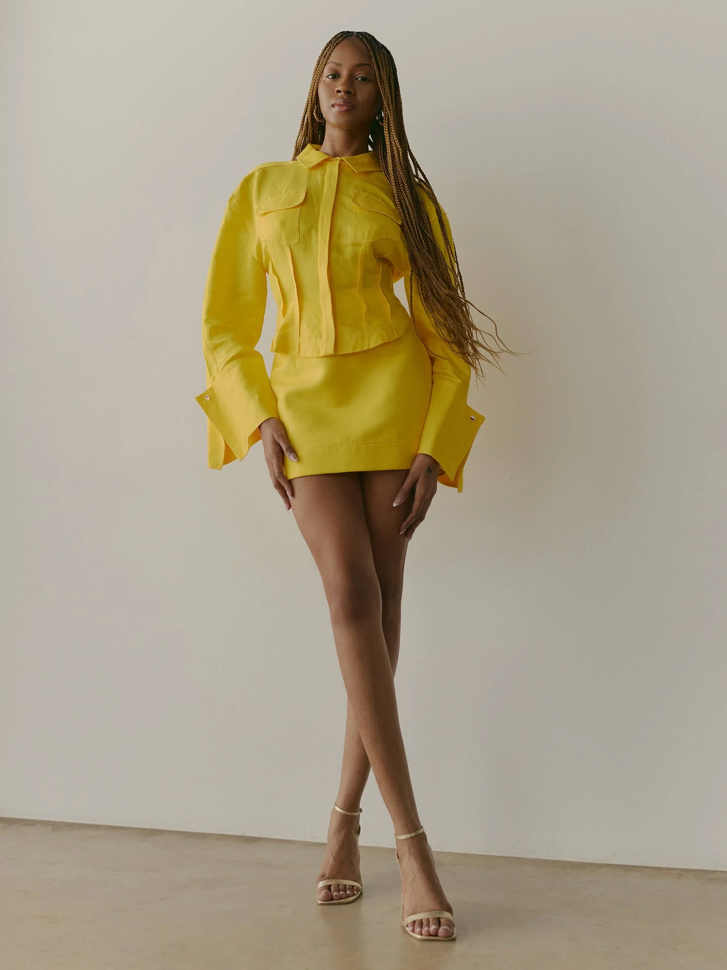A woman stands against a plain white wall, wearing a yellow jacket and skirt, beige high heels, and gold jewelry, with long braids.