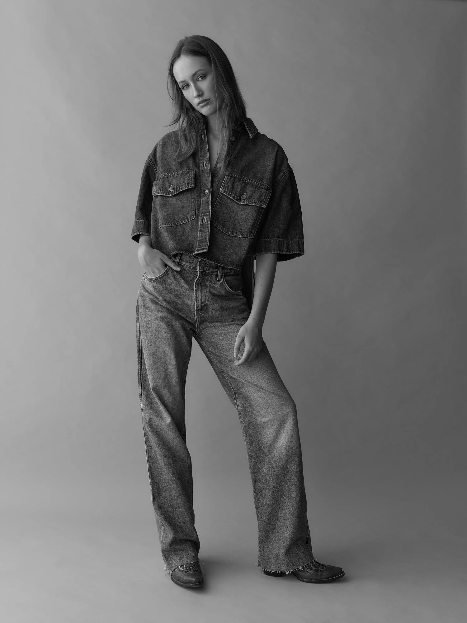 Black and white photo of a woman standing against a plain background, wearing a denim jacket, high-waisted jeans, and black shoes, looking at the camera with a relaxed pose.