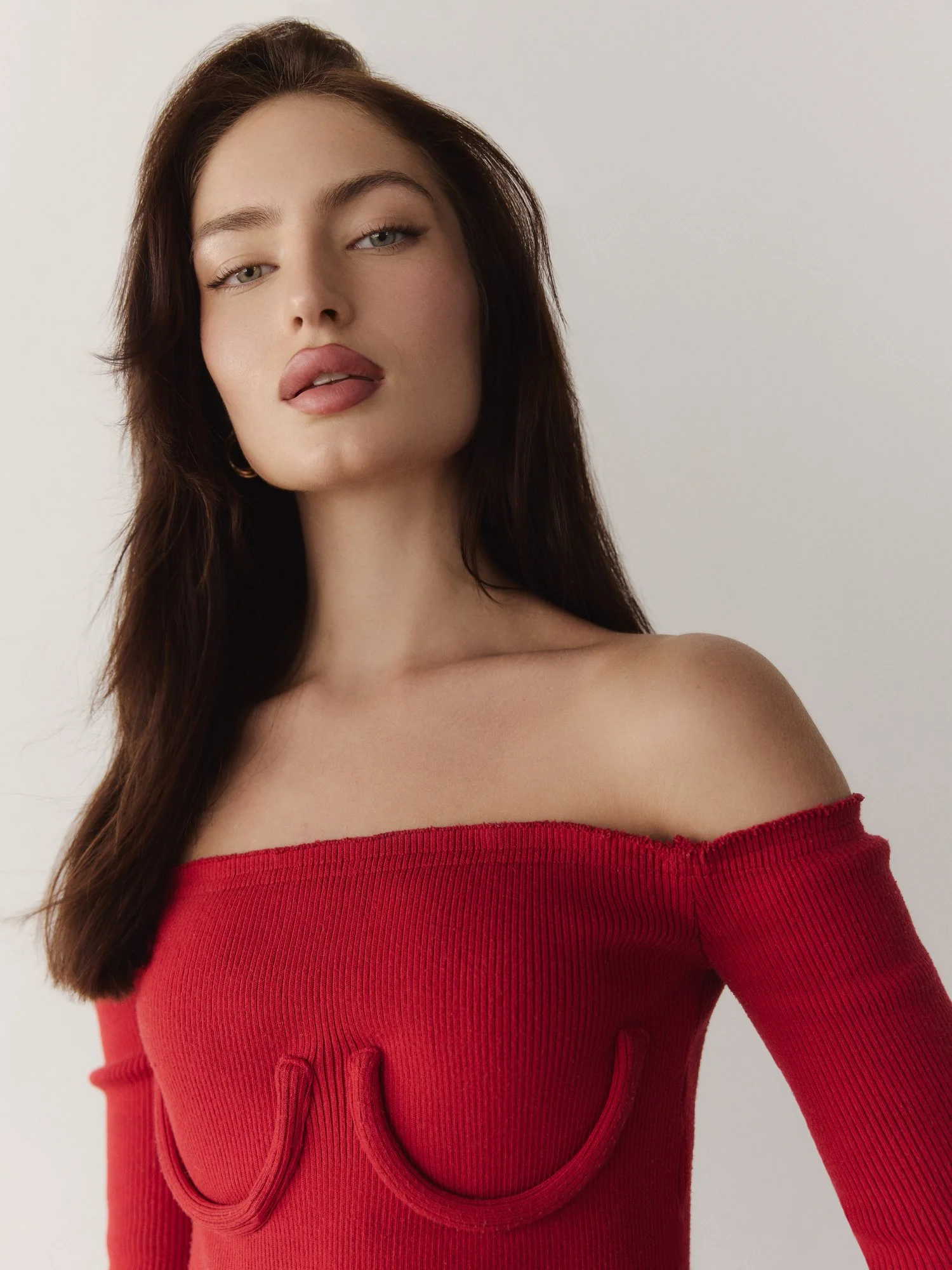 A woman with long dark hair, wearing a red off-the-shoulder top, looking directly at the camera against a plain background.