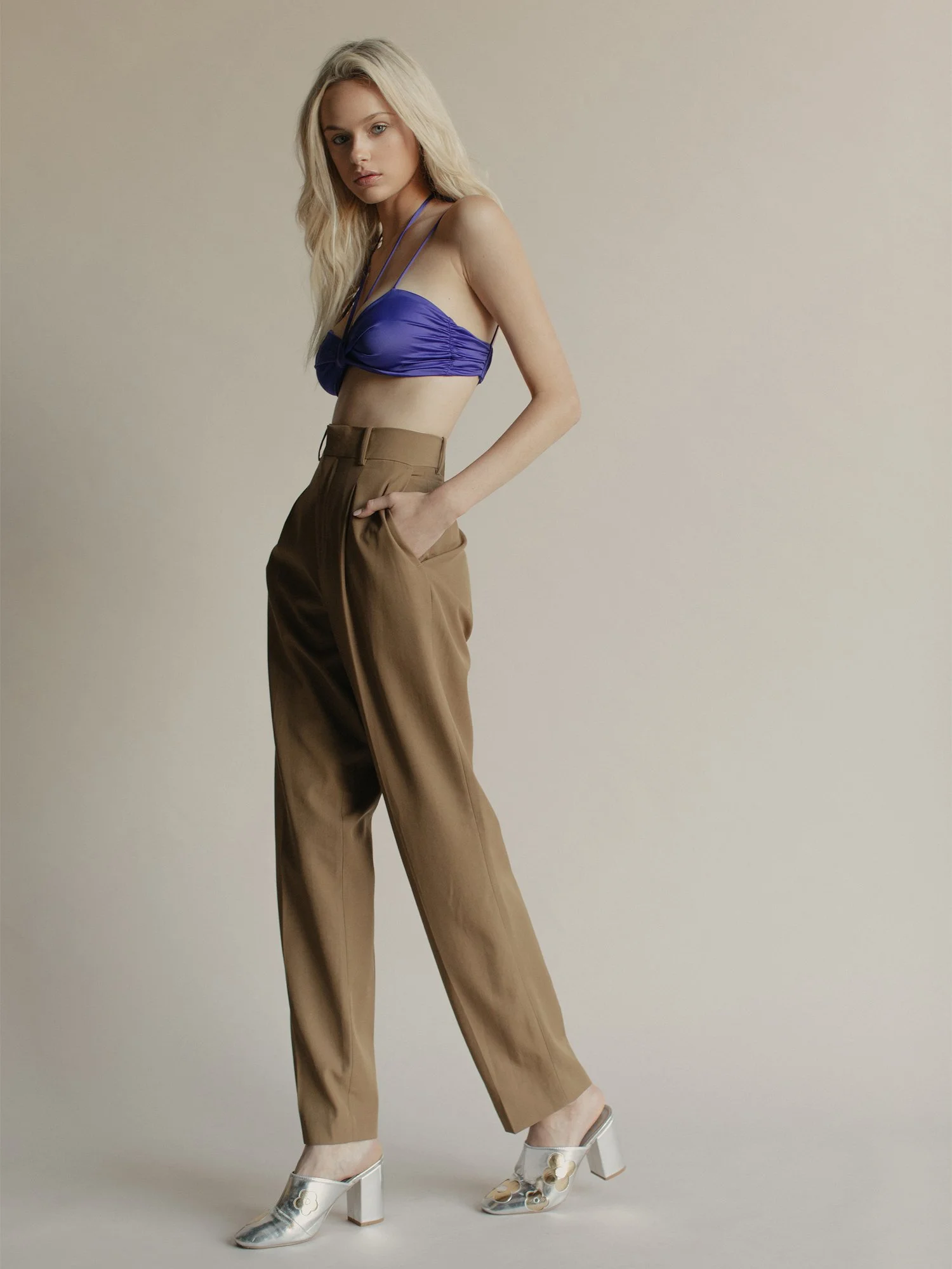 A woman with long blonde hair wearing a purple crop top, tan high-waisted trousers, and transparent high-heeled shoes with floral designs, standing against a neutral background.