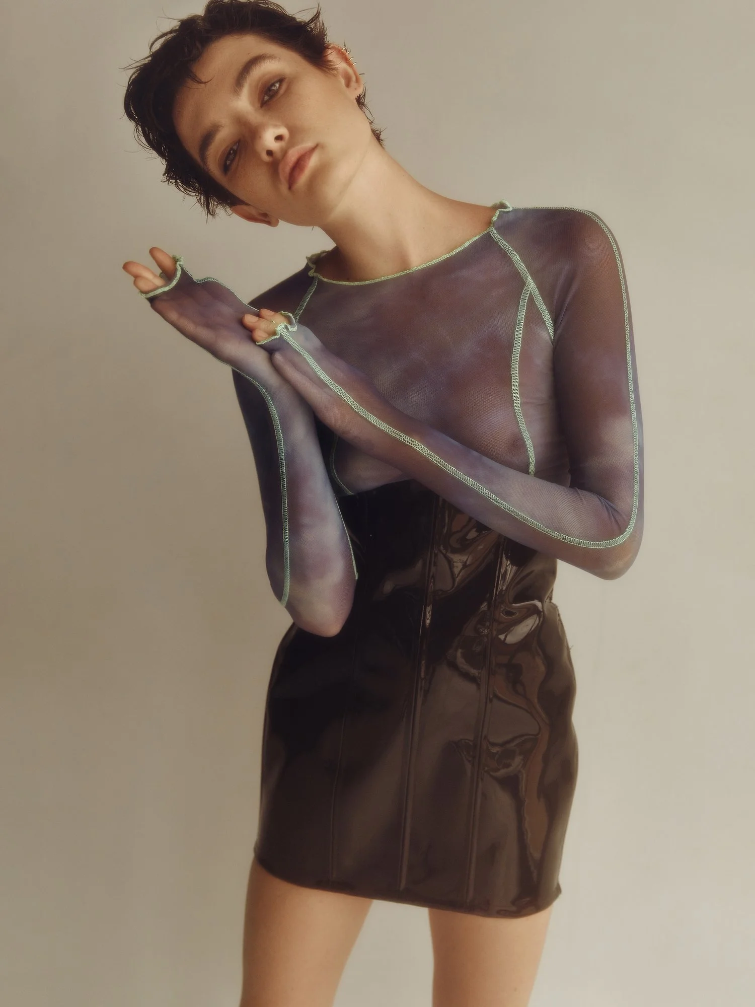 A person with short dark hair has a relaxed expression, slightly tilted head, and is wearing a sheer, long-sleeved, dark top with contrasting green stitching. The person's hand is near their face, and they are wearing a shiny, dark, high-waisted skir