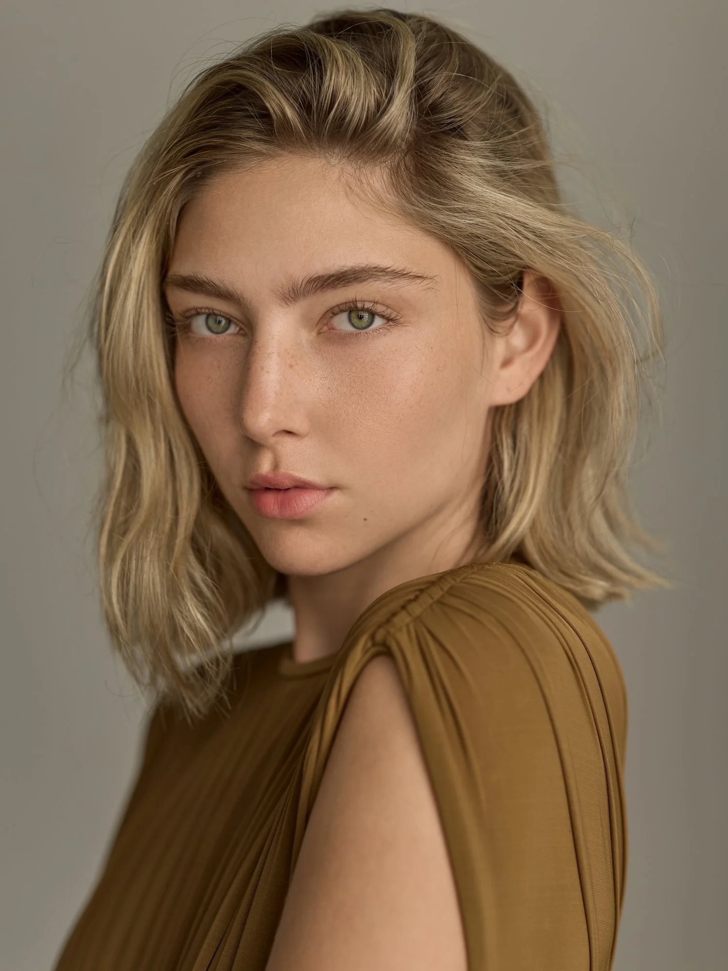 Close-up of a woman with blonde hair and green eyes wearing a brown top against a neutral background.