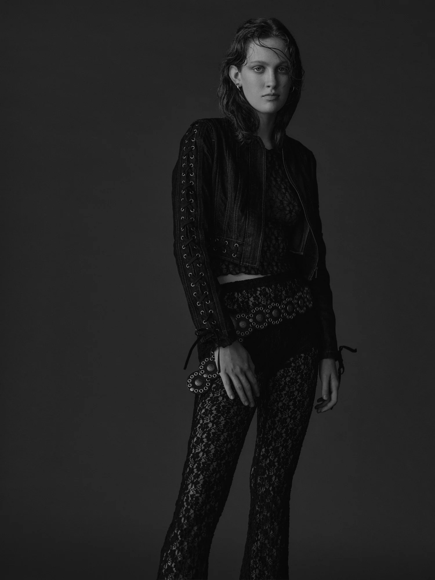 Black and white portrait of a young woman with wet hair, wearing a lace top, a leather jacket with lace-up sleeves, and patterned lace pants, against a dark background.
