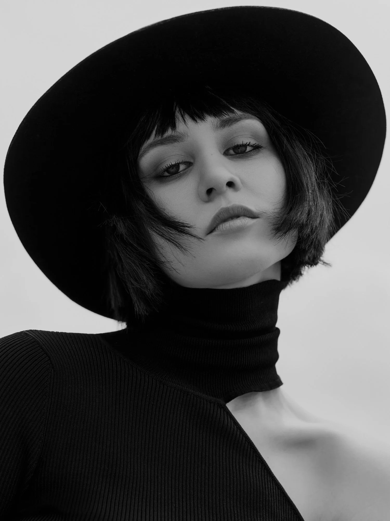 A black-and-white close-up portrait of a woman wearing a large wide-brimmed hat and a ribbed turtleneck.
