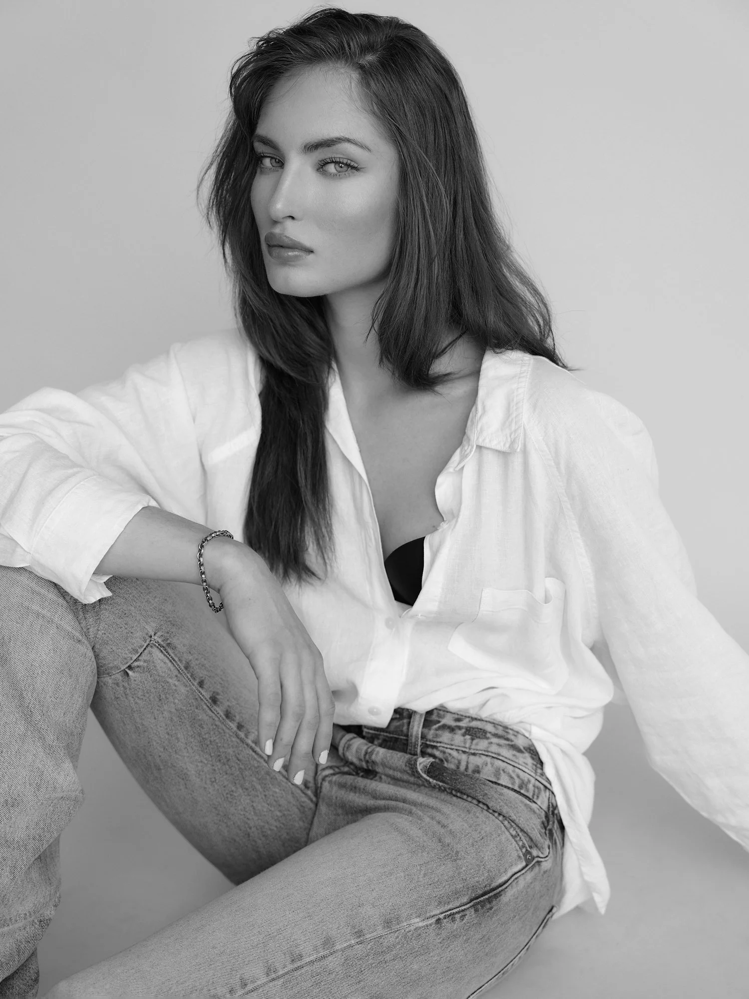 Black and white photo of a woman with long hair, wearing a white shirt and jeans, sitting with one knee up, against a plain background.