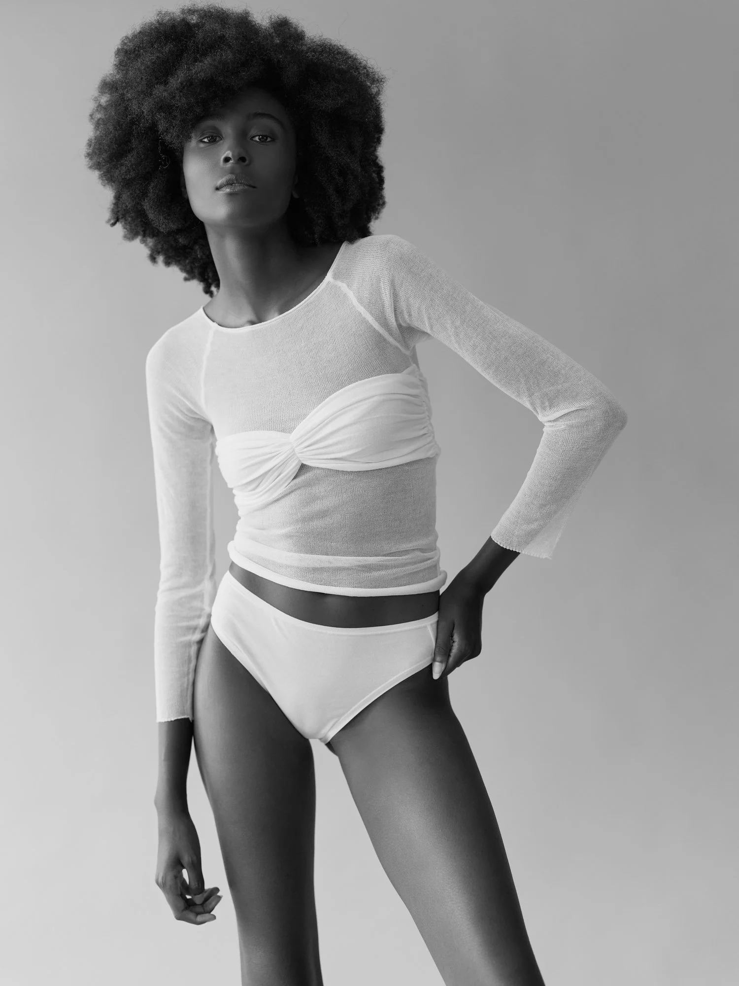 A black and white photo of a woman with curly hair, wearing a long-sleeve top with a bow detail on the front, and high-waisted shorts, standing against a plain background.