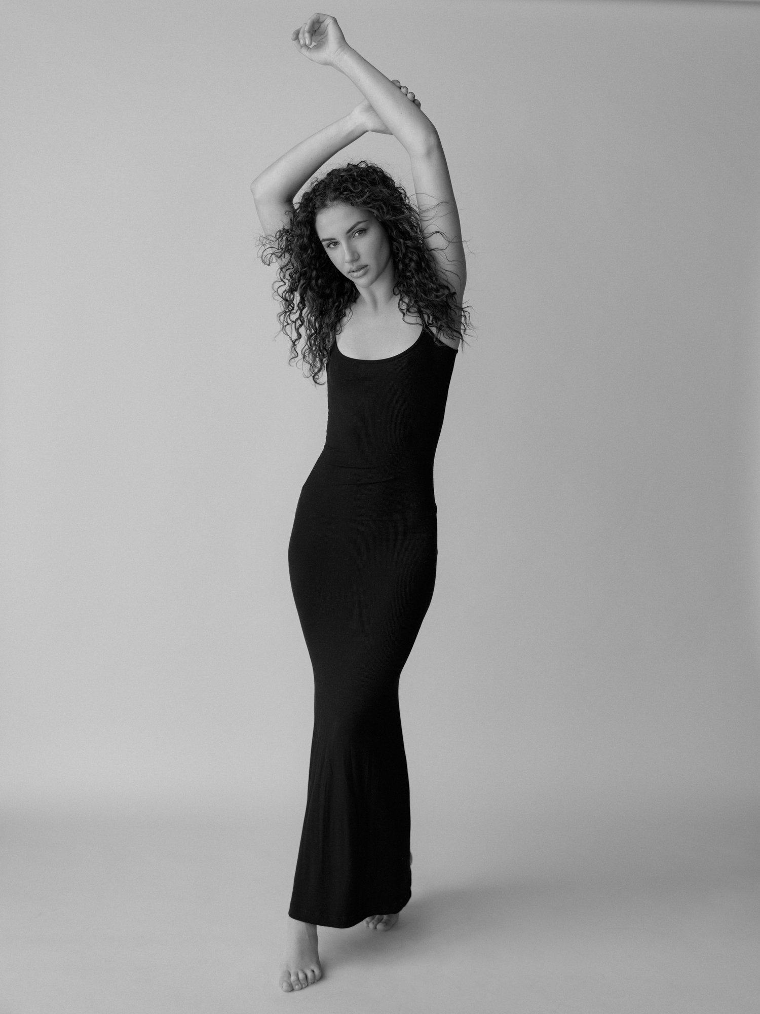 A black-and-white photo of a woman with curly hair wearing a long black dress, standing with one leg slightly forward and holding her arms above and behind her head in a dance pose, against a plain background.