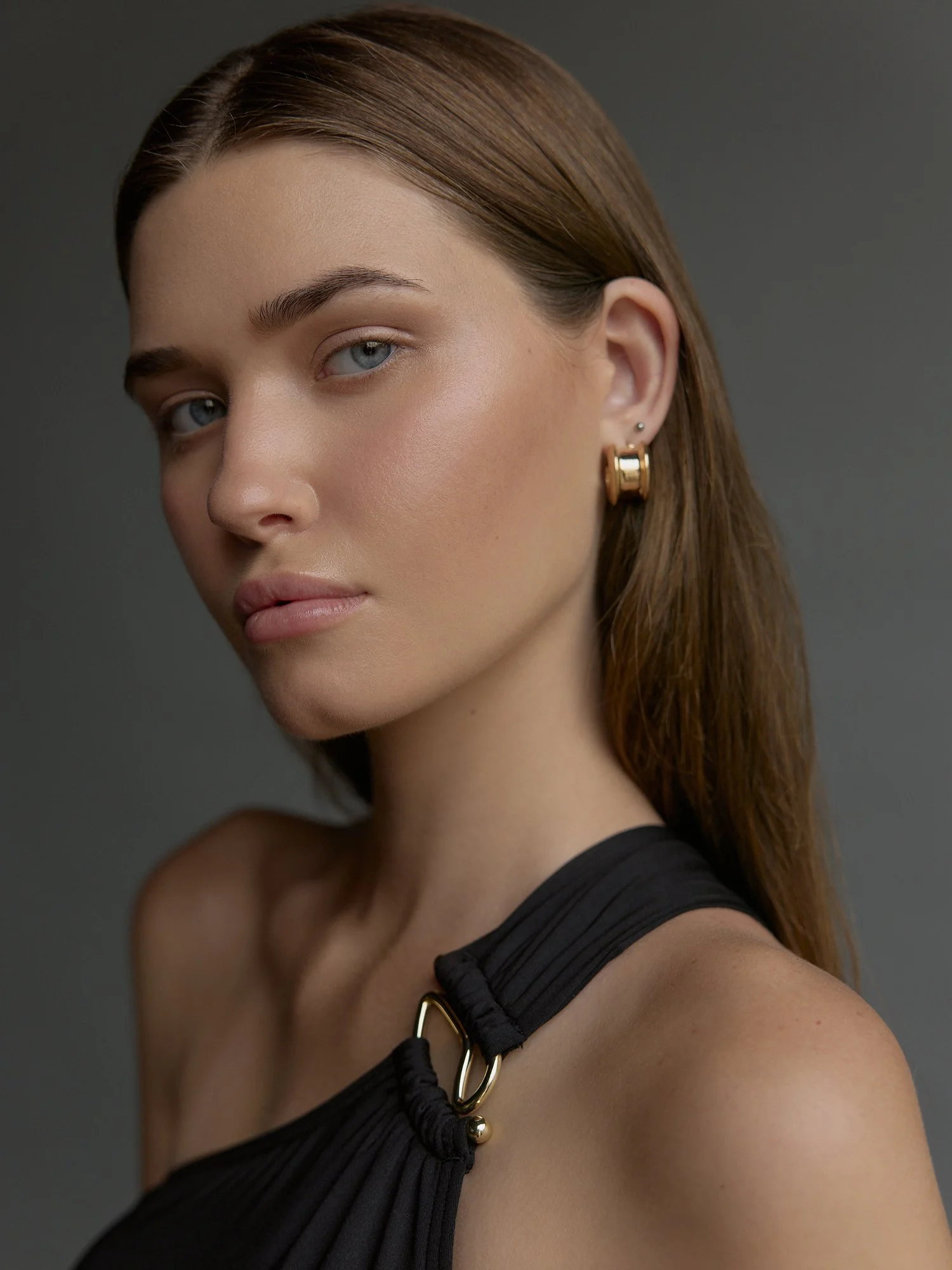 Close-up of a woman with brown hair, light eye makeup, wearing a black top and gold earrings, posing against a dark background.