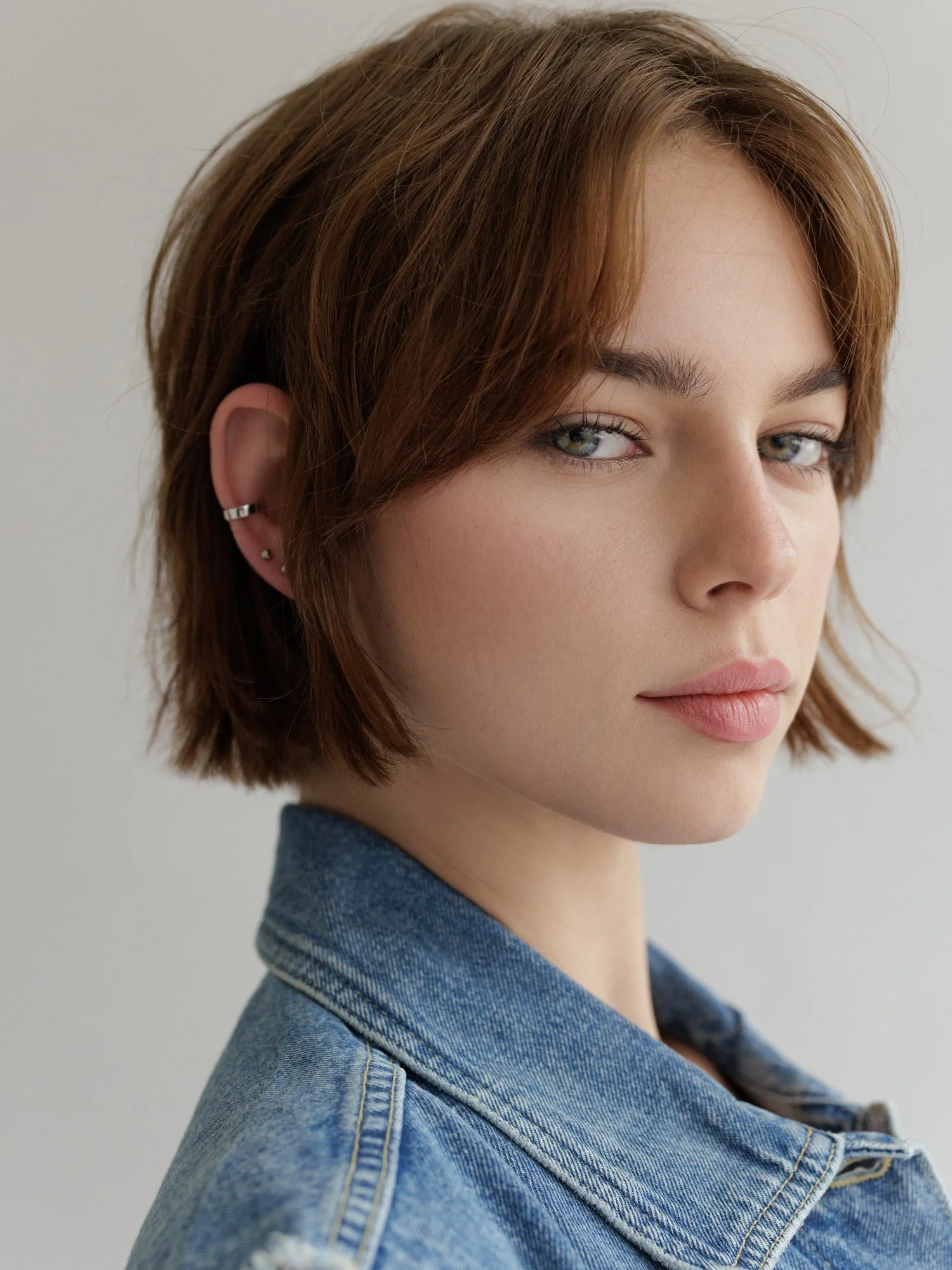 Close-up of a young woman with short auburn hair, green eyes, light makeup, wearing a blue denim jacket, looking at the camera.