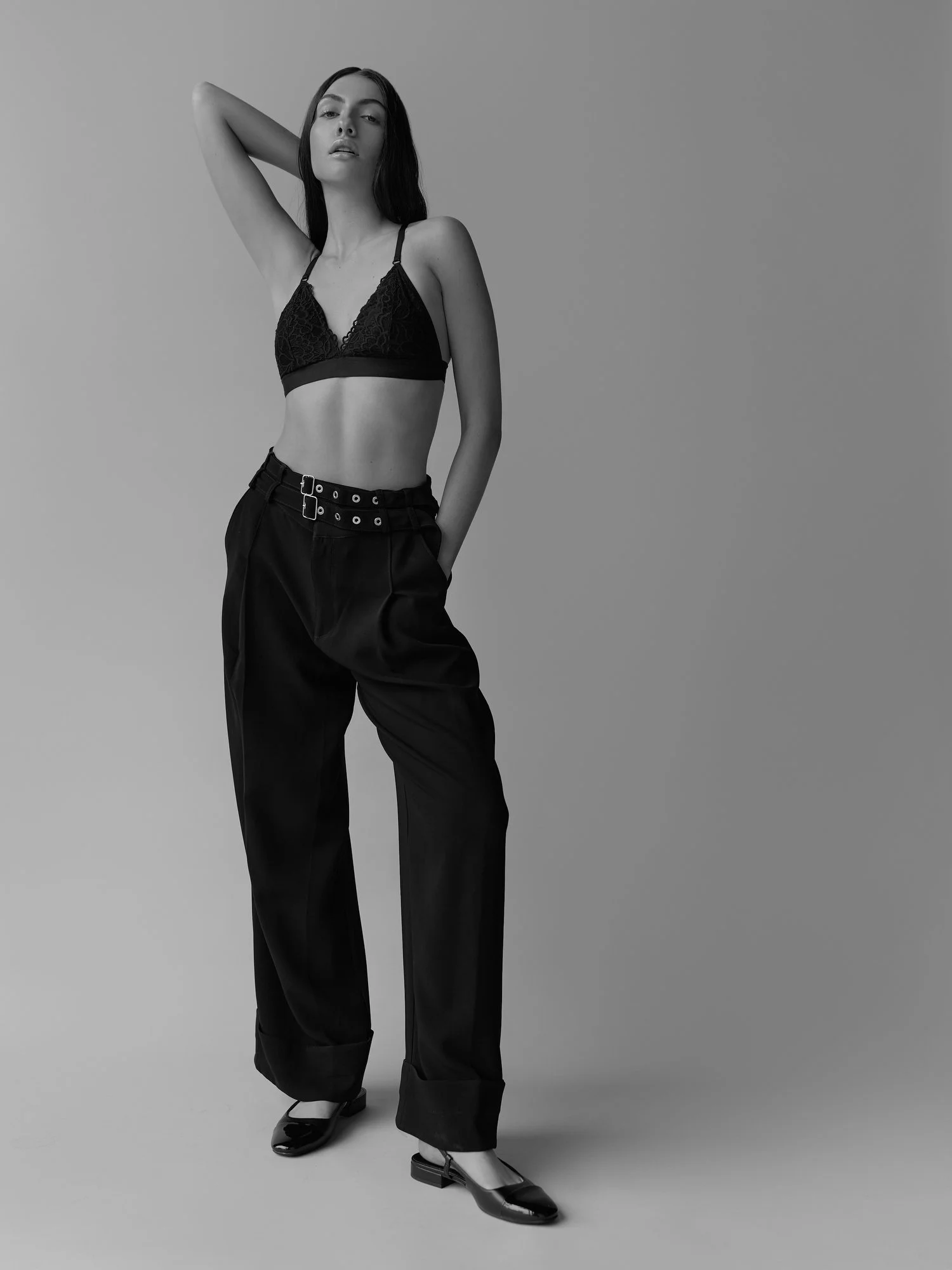 A young woman with long dark hair wearing a black lace bralette top, black high-waisted pants, and shiny black shoes with a low heel, standing with one arm raised behind her head against a plain gray background.