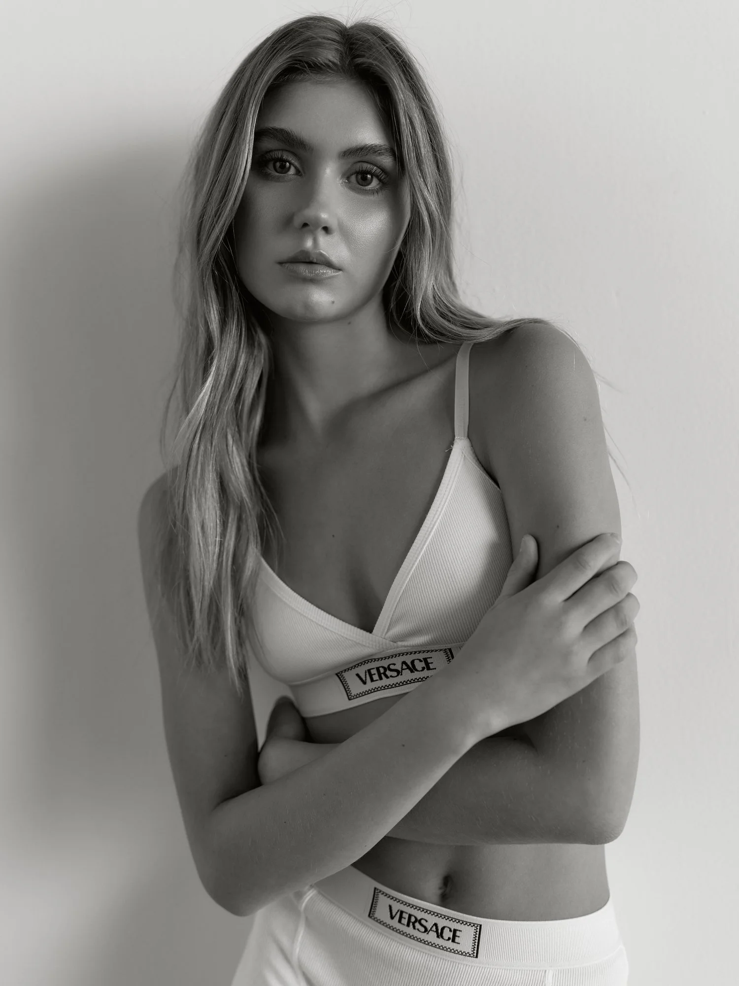 Black-and-white photo of a young woman wearing a Versace sports bra and matching bottoms, with long wavy hair, crossed arms, and looking directly at the camera against a plain background.