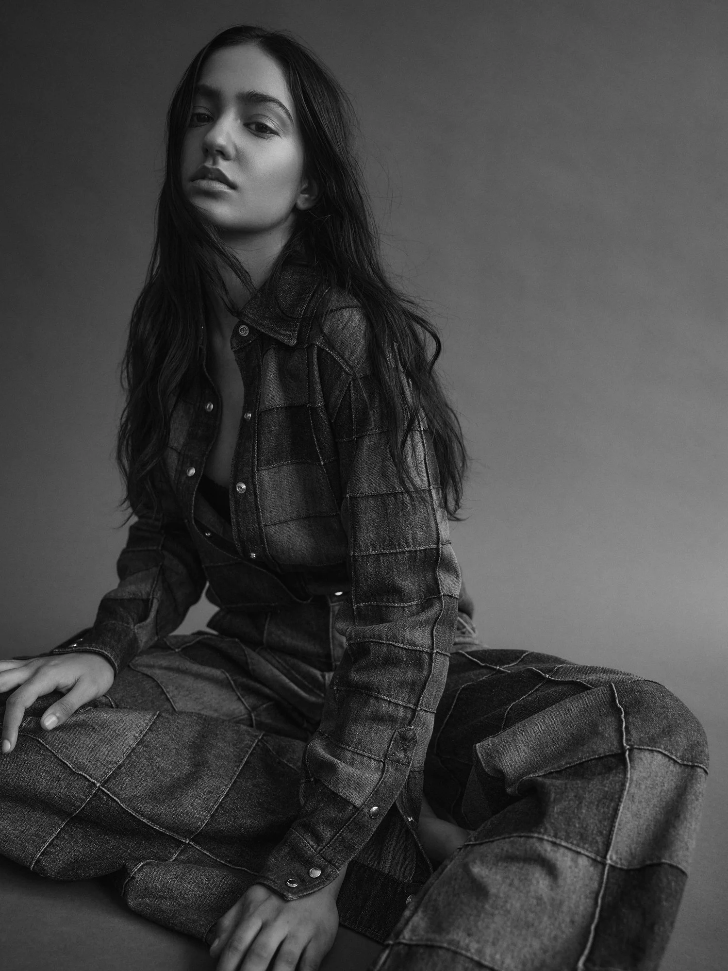 Black and white photo of a woman in denim jacket and jeans sitting on the floor, looking at the camera with a neutral expression.