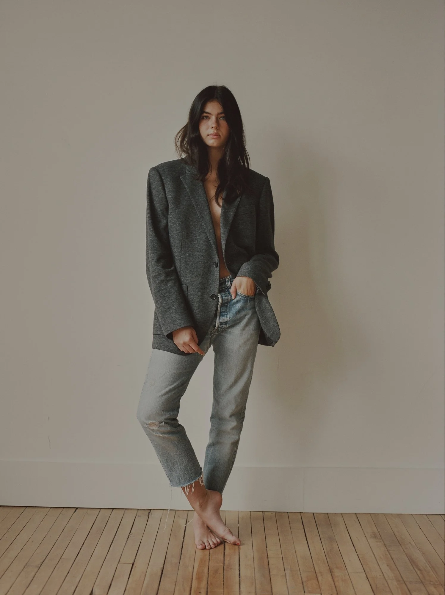 A woman with dark hair wearing a gray blazer and distressed jeans standing barefoot on a wooden floor against a plain wall.