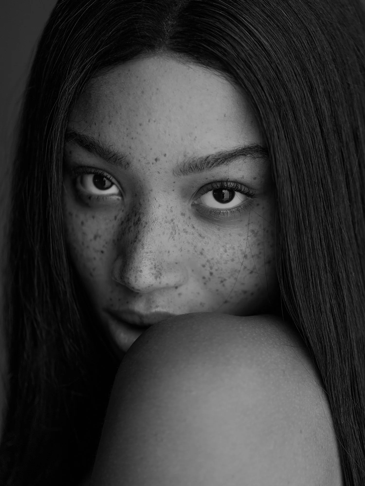 Close-up black and white photo of a woman's face, showing her eyes, eyebrows, and forehead, with long hair partially covering her face, and her resting her chin on her arm.