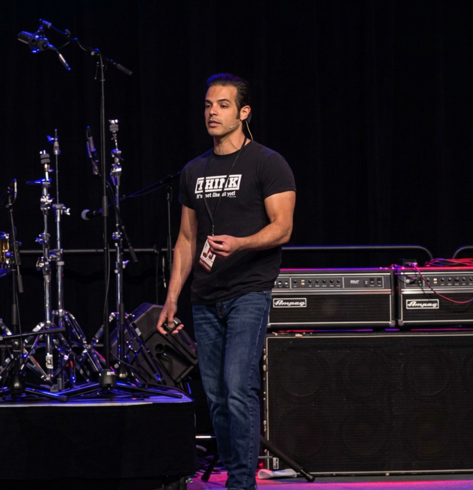 J Gulinello HRP Co Founder wearing a black t-shirt with white text, holding a microphone, standing on a stage with musical equipment, including drum kits and amplifiers, behind him.