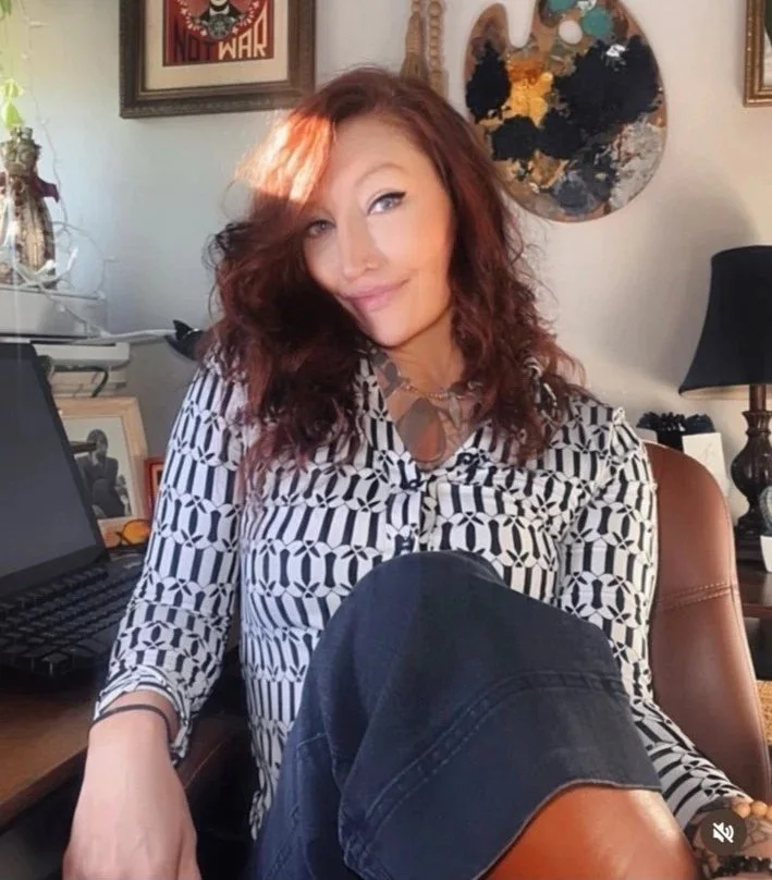 Jody Barna CTO bio Picture in her design studio wearing a patterned black and white blouse and black pants, sitting in a cozy room with artwork on the walls and a lamp in the background.