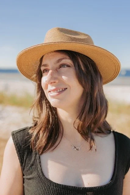 Woman wearing a wide-brimmed straw hat and a black sleeveless top, smiling outdoors on a sunny day at the beach or seaside.